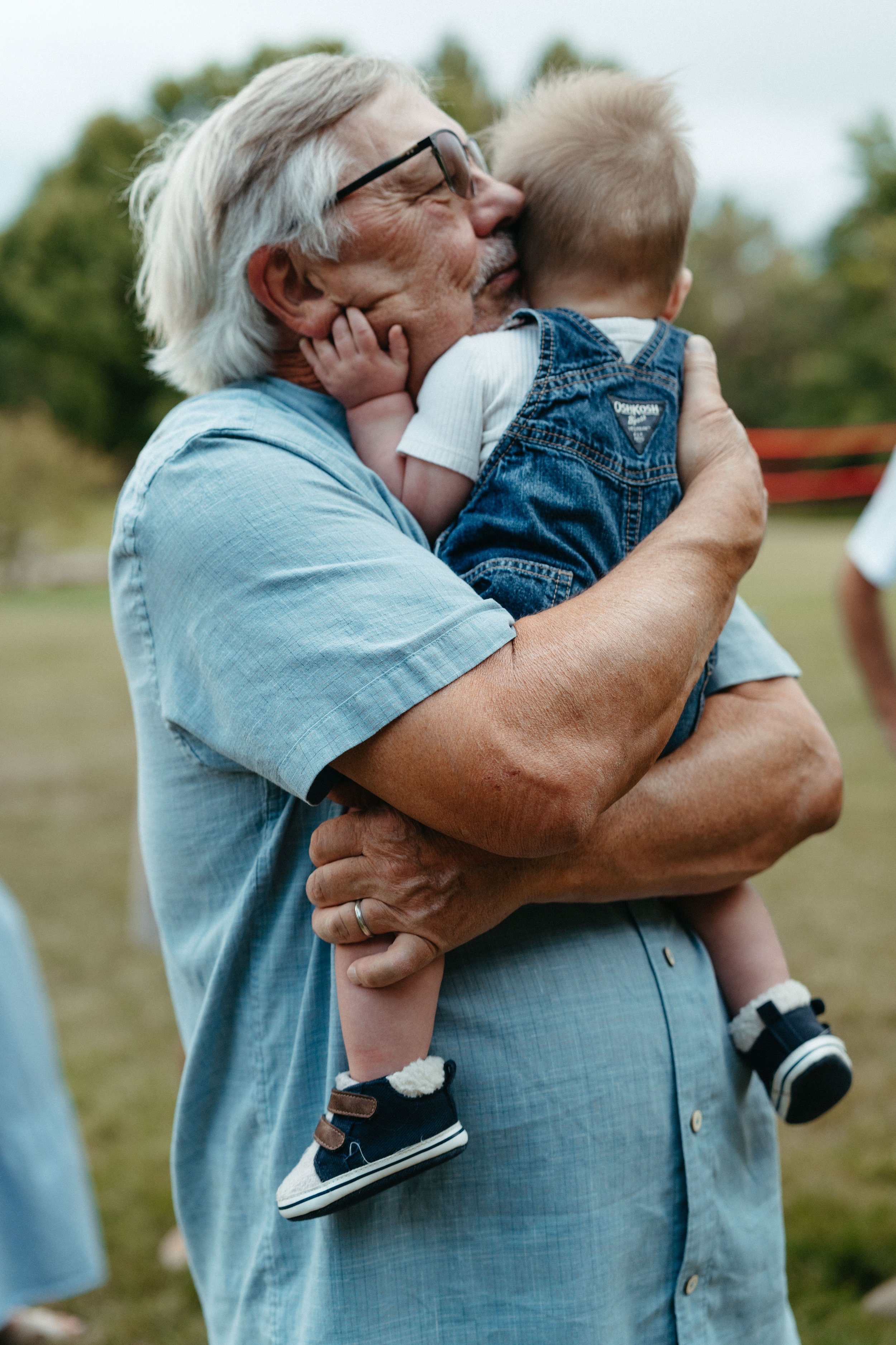 An elderly man with gray hair, glasses, and a light blue shirt holding a young boy with blonde hair and a denim vest, outdoors in a park or open area.
