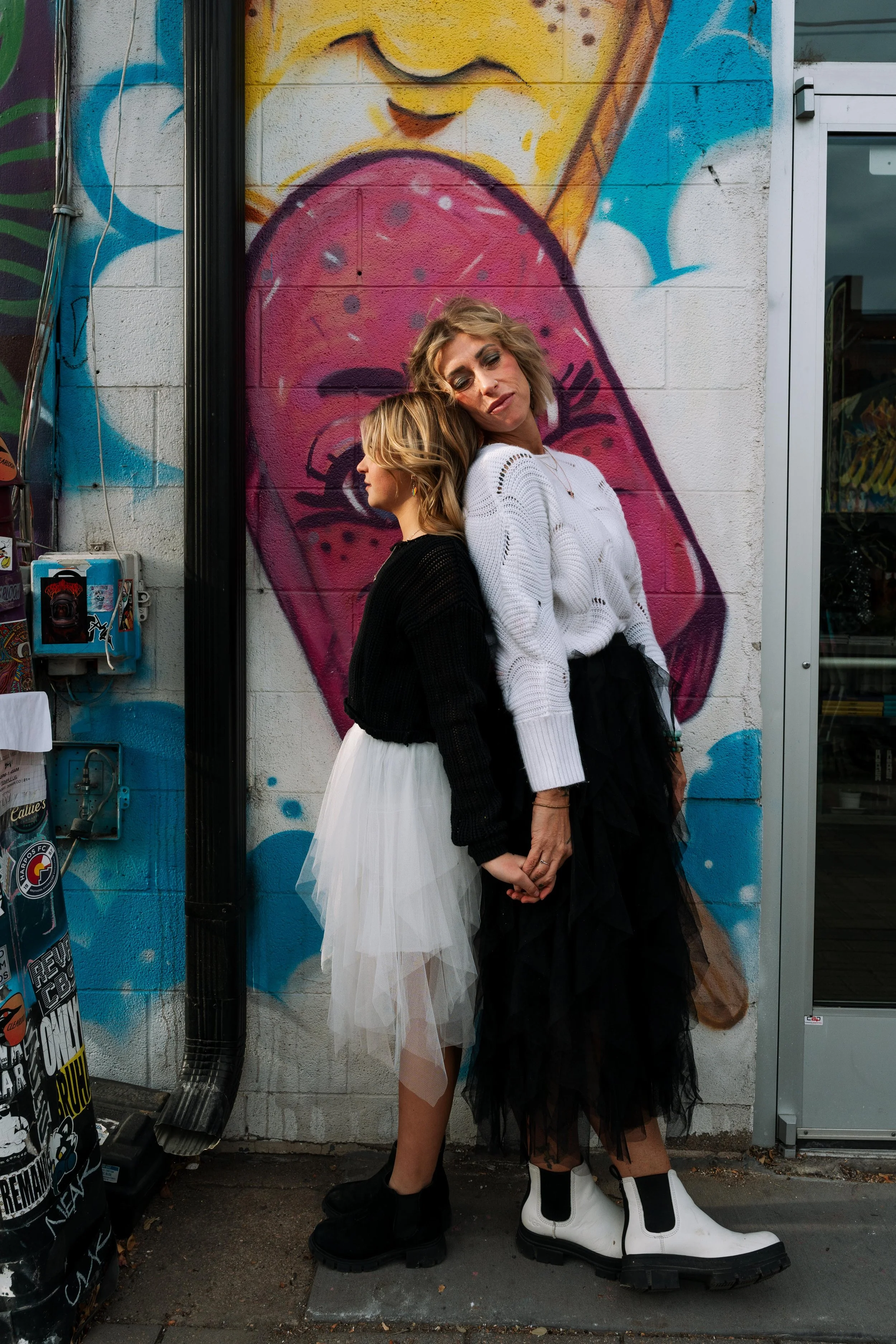 Two women standing back to back holding hands, standing in front of a colorful mural of a woman's face with her tongue out.