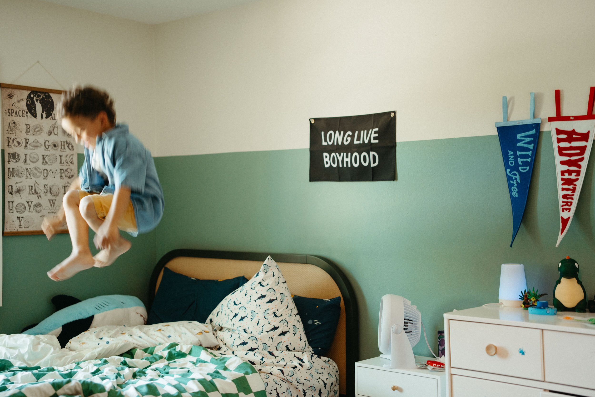 A boy is jumping on a bed in a bedroom with a green and white geometric duvet cover, and dinosaur-patterned pillows. The room has a black calendar or poster and a freestanding fan on a white dresser, with toys and decorations. There are signs on the 