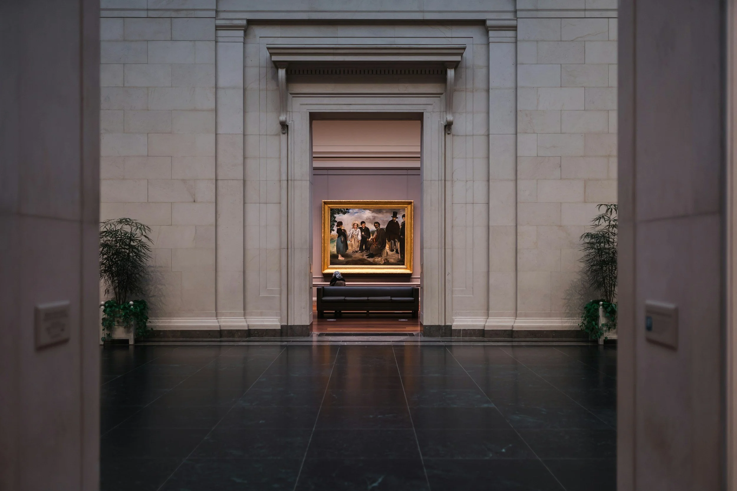 Museum interior with a large framed painting on the wall and a black bench in front, flanked by potted plants.