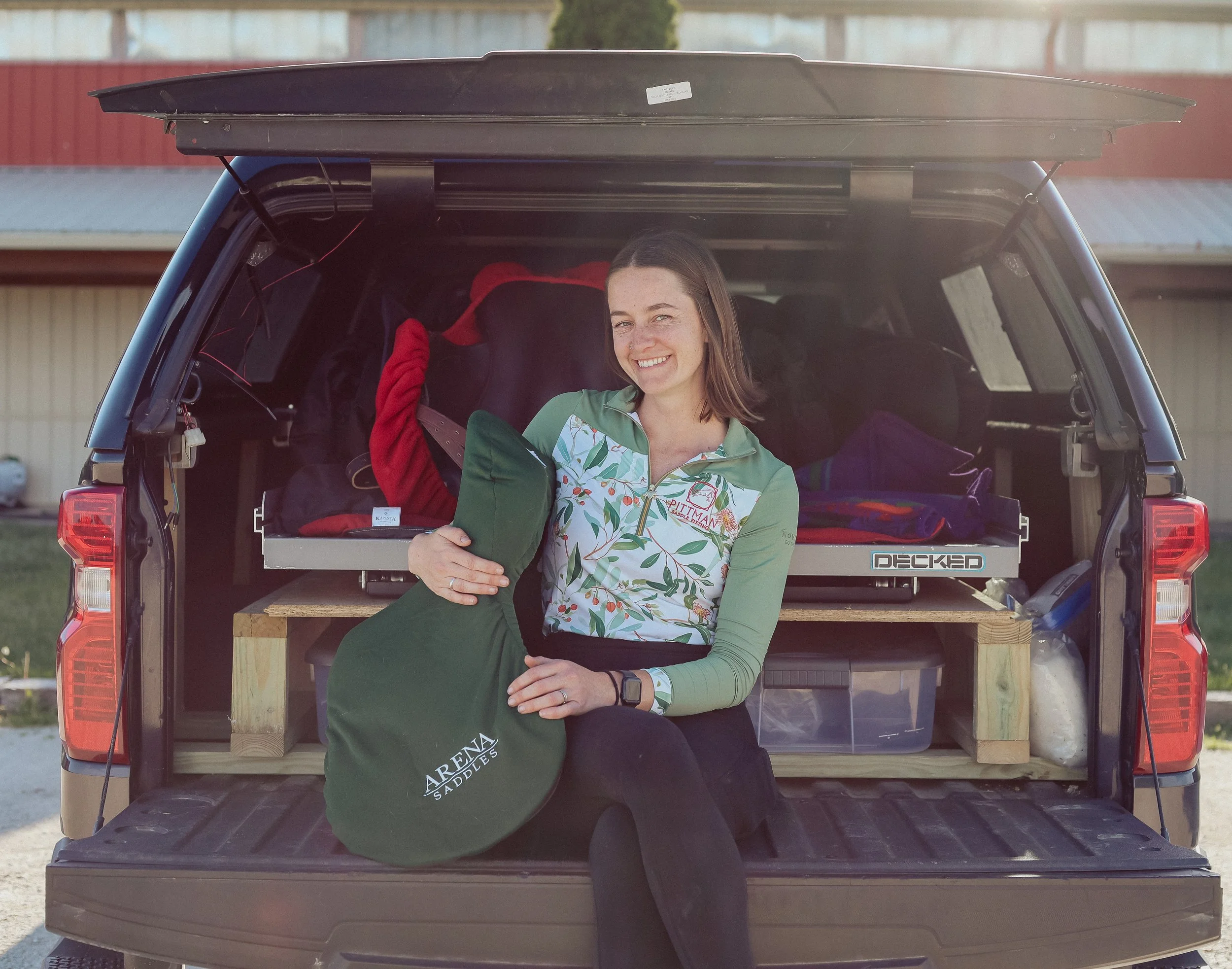 A woman smiling and holding a paddle, sitting inside the open tailgate of a pickup truck loaded with gear, at an outdoor location with a building in the background.