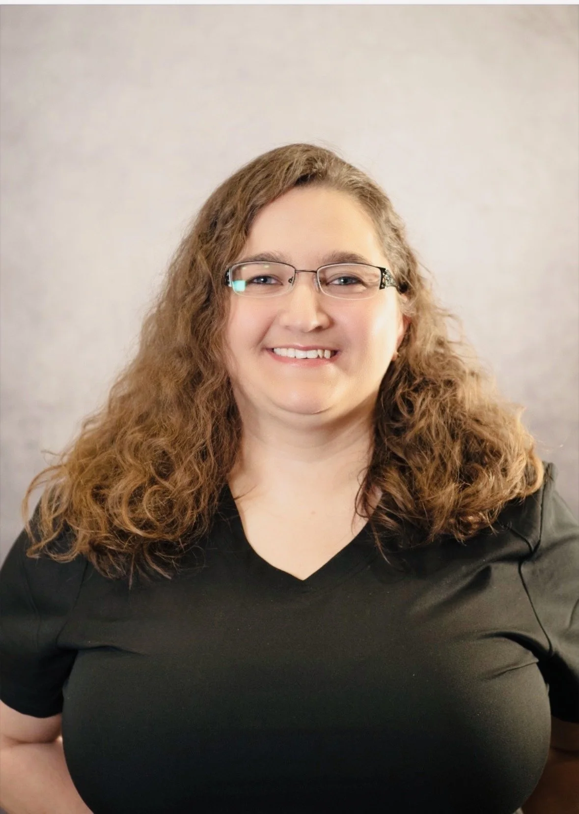 Person with curly hair and glasses, wearing a black "Massage Envy" shirt, smiling against a neutral background.