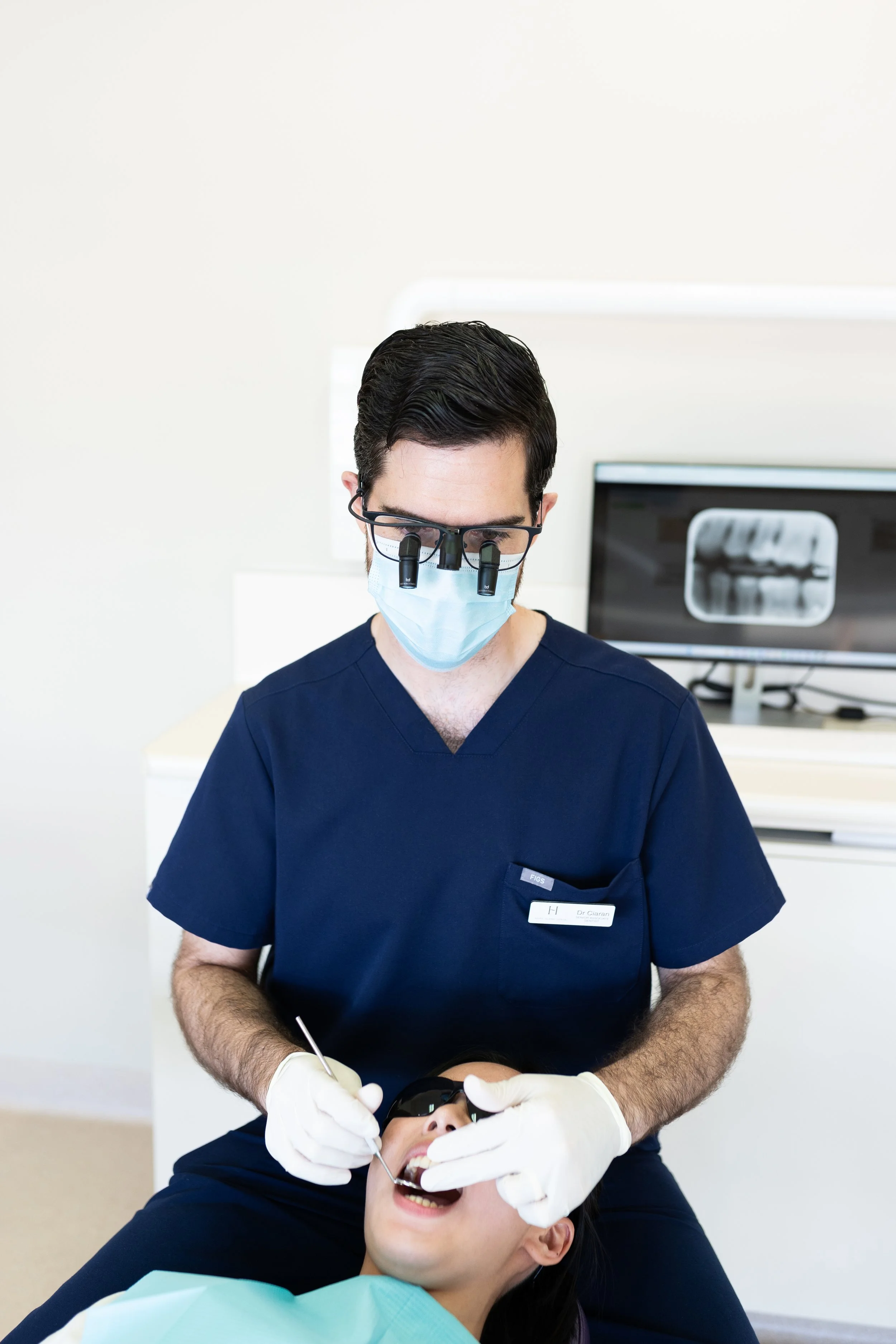 Associate dentist Dr Ciaran Charalambou at Hope Island Dental wearing loupes, a face mask and a navy scrubs examining a patient.