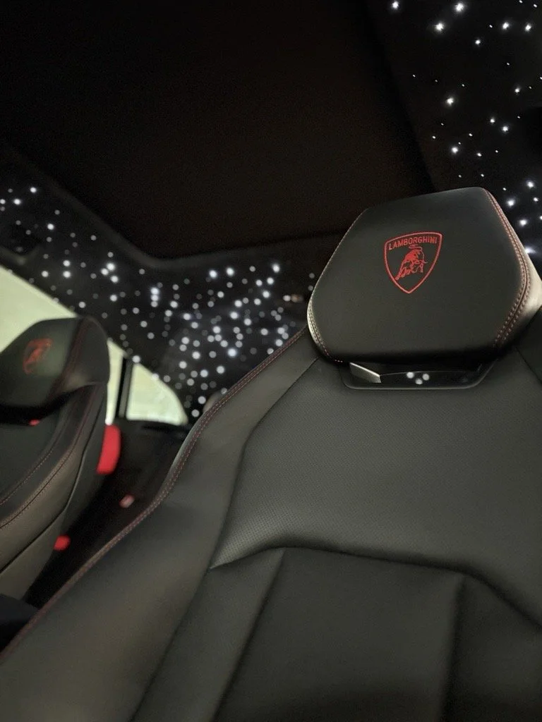 The interior of a luxury Lamborghini vehicle featuring black leather seats with red stitching and the Lamborghini logo, a starry night sky-style ceiling with illuminated dots.