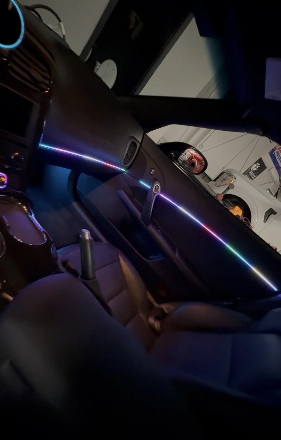 Interior of a car with a dashboard featuring colorful LED lights, a steering wheel, and front seats in a dark garage with a white vehicle parked outside.