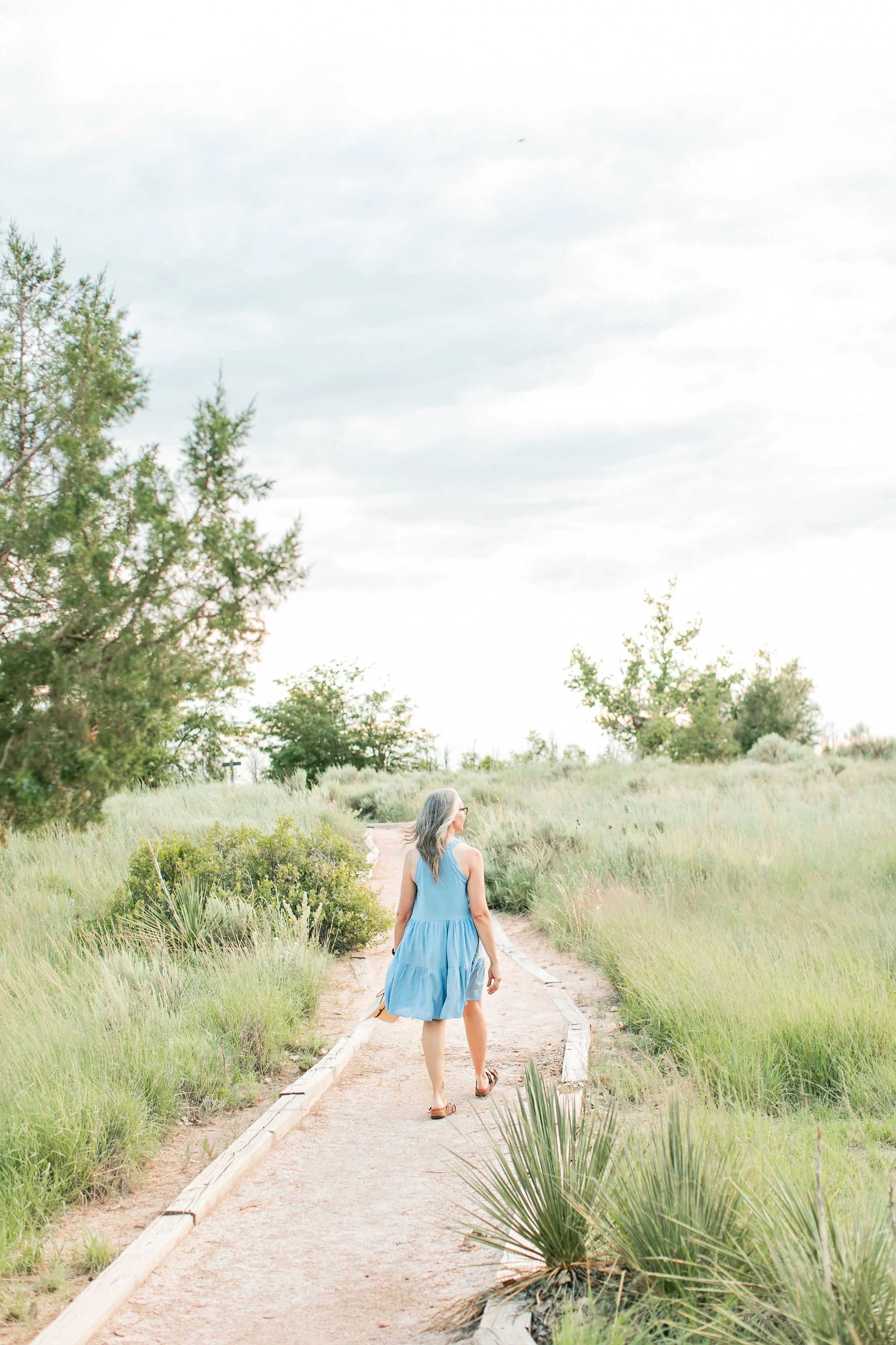 A woman in a blue dress walking on a sandy path through a grassy nature area with trees.