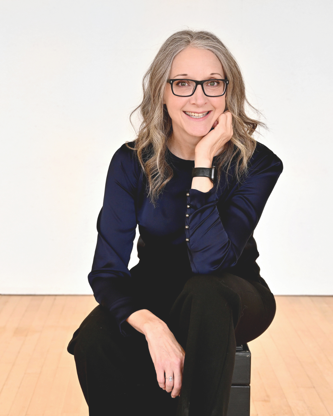 A woman with gray hair wearing glasses and a navy blue satin blouse, sitting on a black box with a wooden floor and white background.