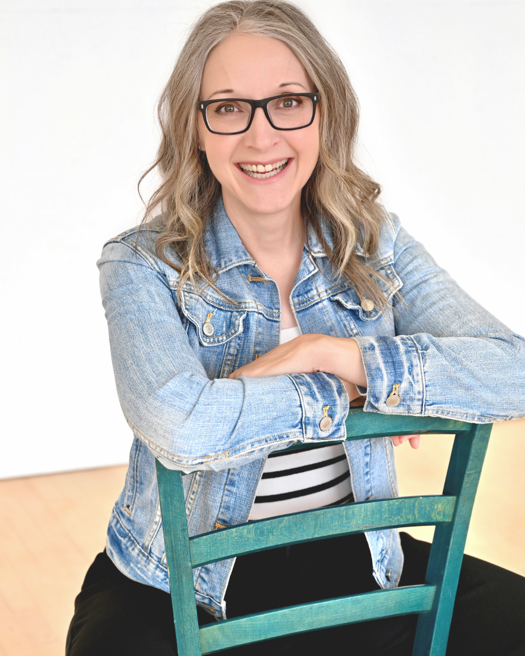 A woman with shoulder-length wavy gray hair wearing black glasses, a denim jacket, and a striped shirt, smiling while sitting on a teal wooden chair against a plain white background.