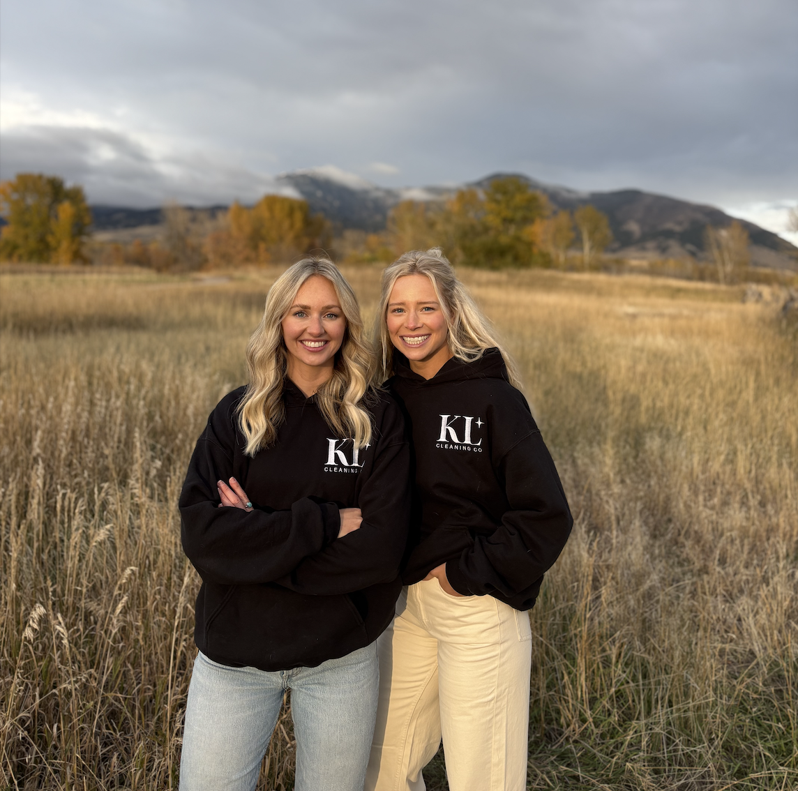 Two young women with blonde hair smiling and standing in a field of tall grass, wearing black hoodies with 'KL Cleaning Co' logo, with mountains and trees in the background on a cloudy day.