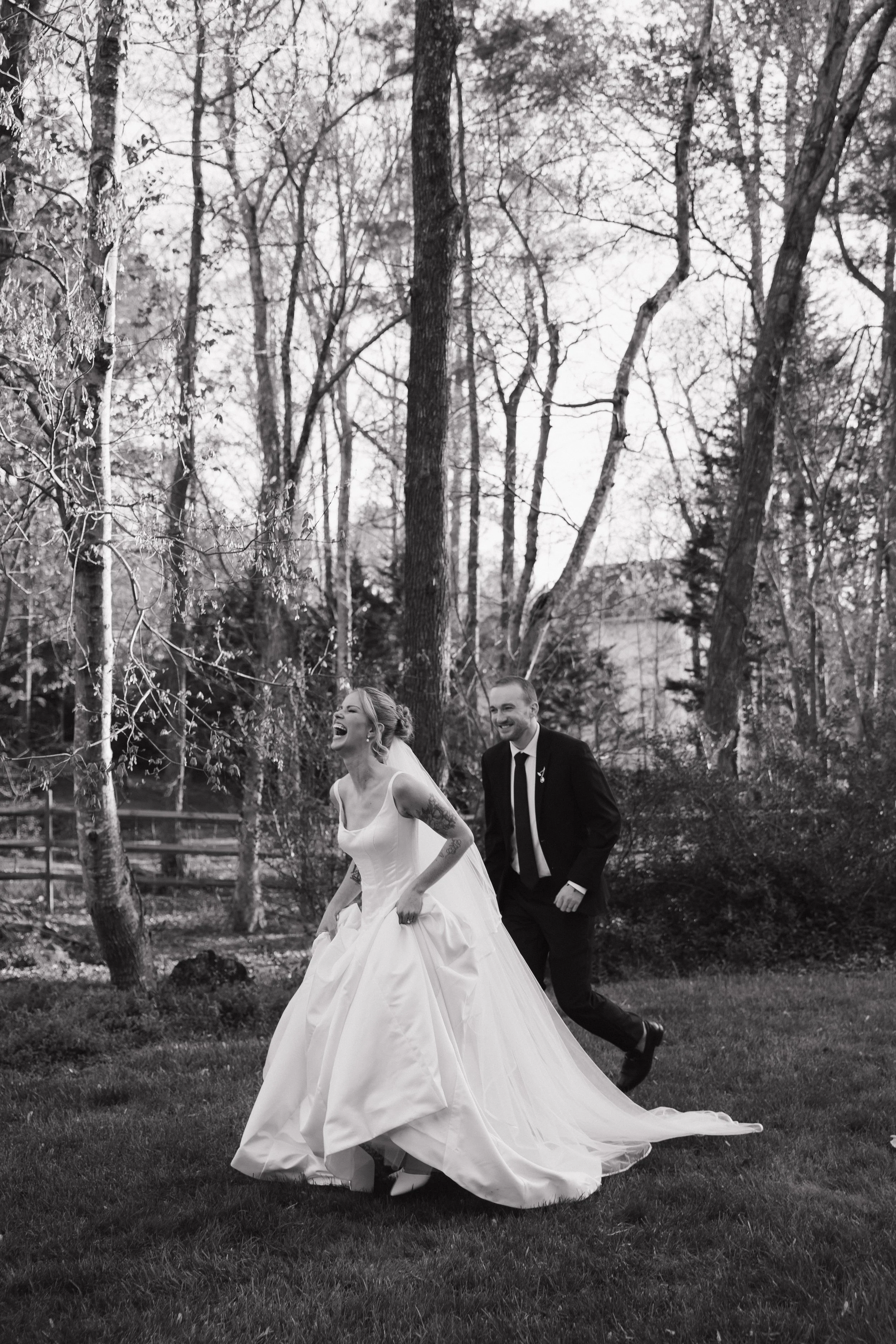 A bride in a white wedding gown laughing and a groom in a black suit with a tie, running in a park with tall trees and bushes in the background.