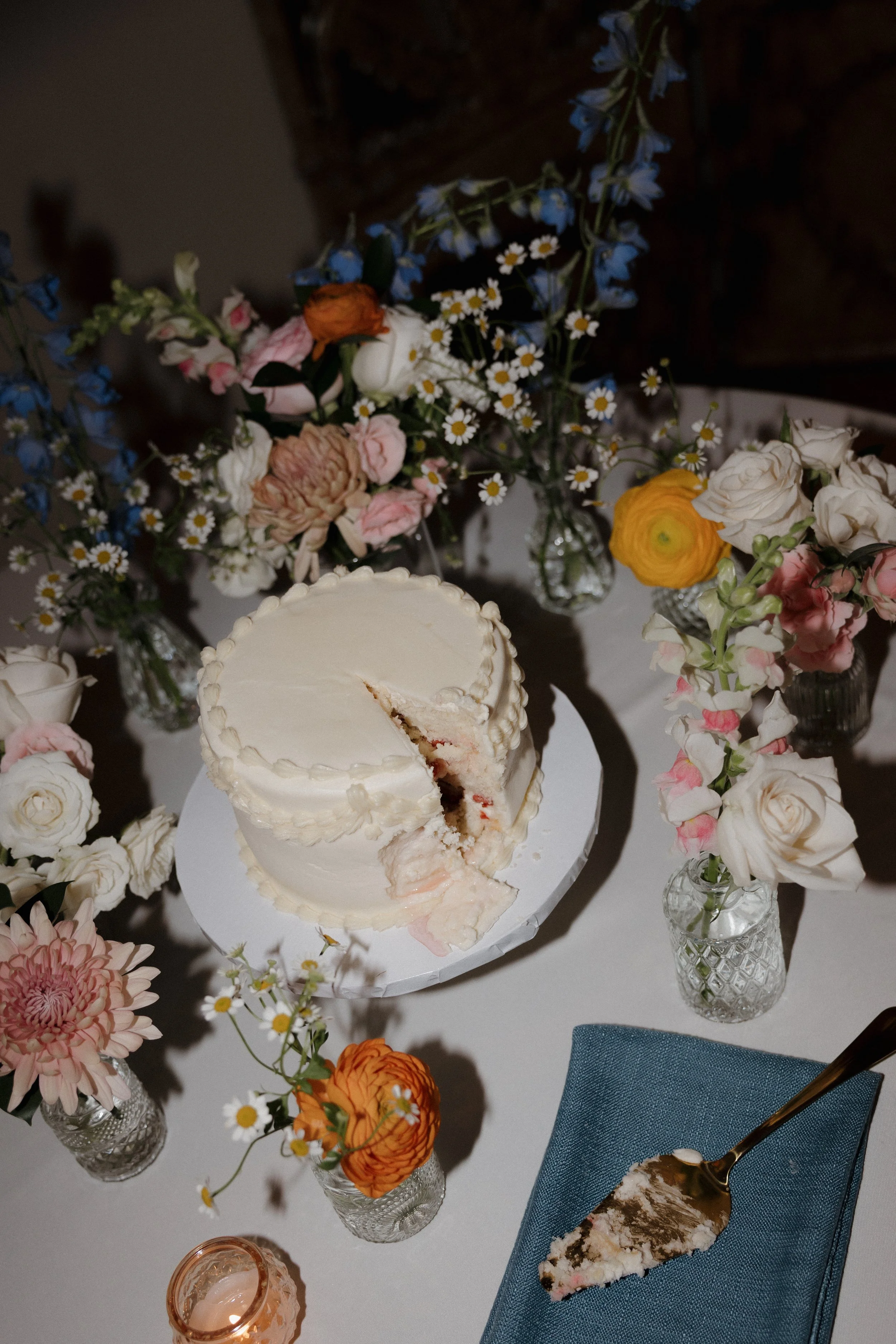 A partially eaten white frosted cake on a white table surrounded by small glass jars filled with pink, orange, yellow, and white flowers in vases, with a blue napkin and fork nearby.