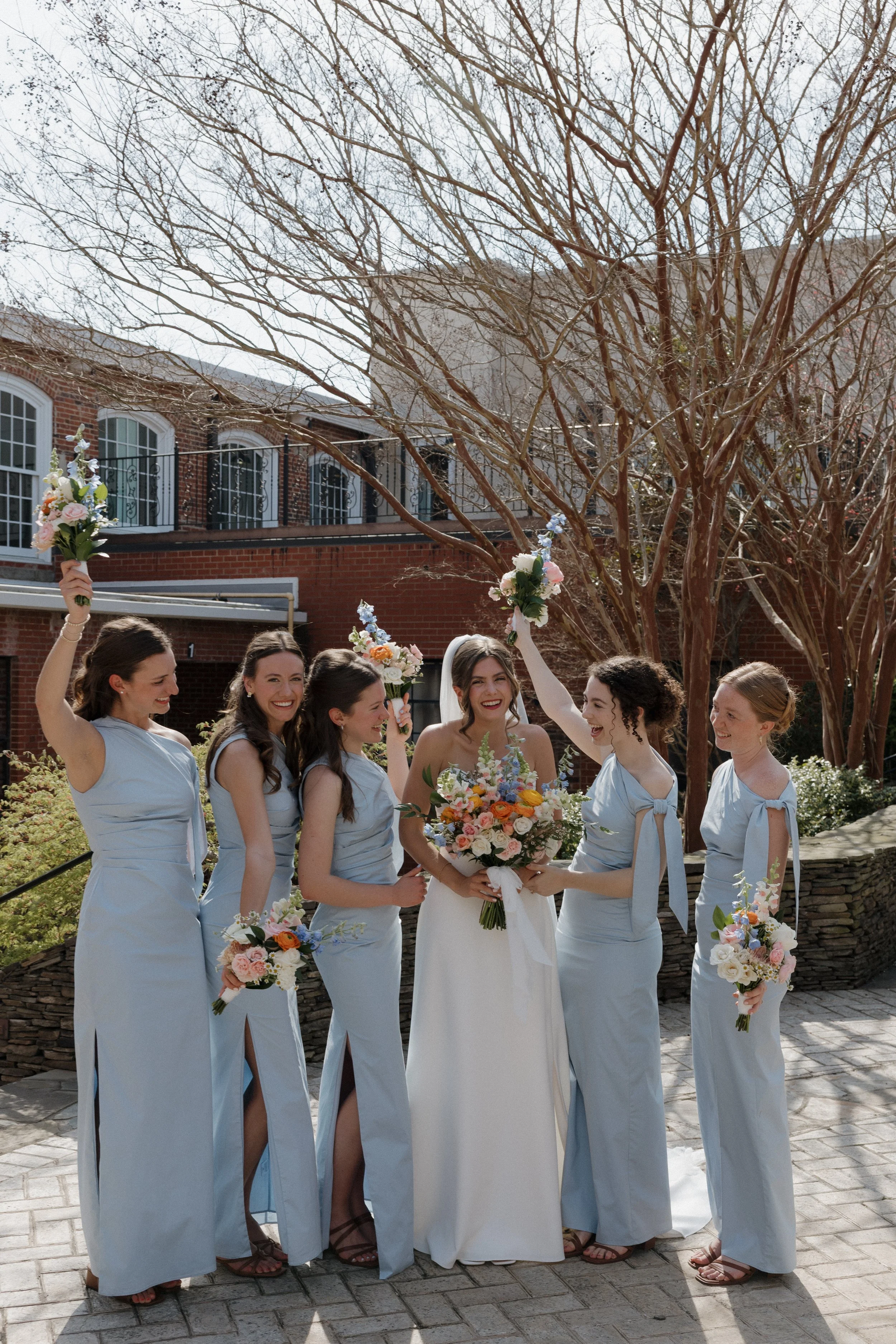 A group of women, likely bridesmaids, celebrating with a bride outdoors during daytime. The bride is holding a large bouquet, and the bridesmaids have smaller bouquets. They are smiling and raising their bouquets in the air, wearing light blue dresse