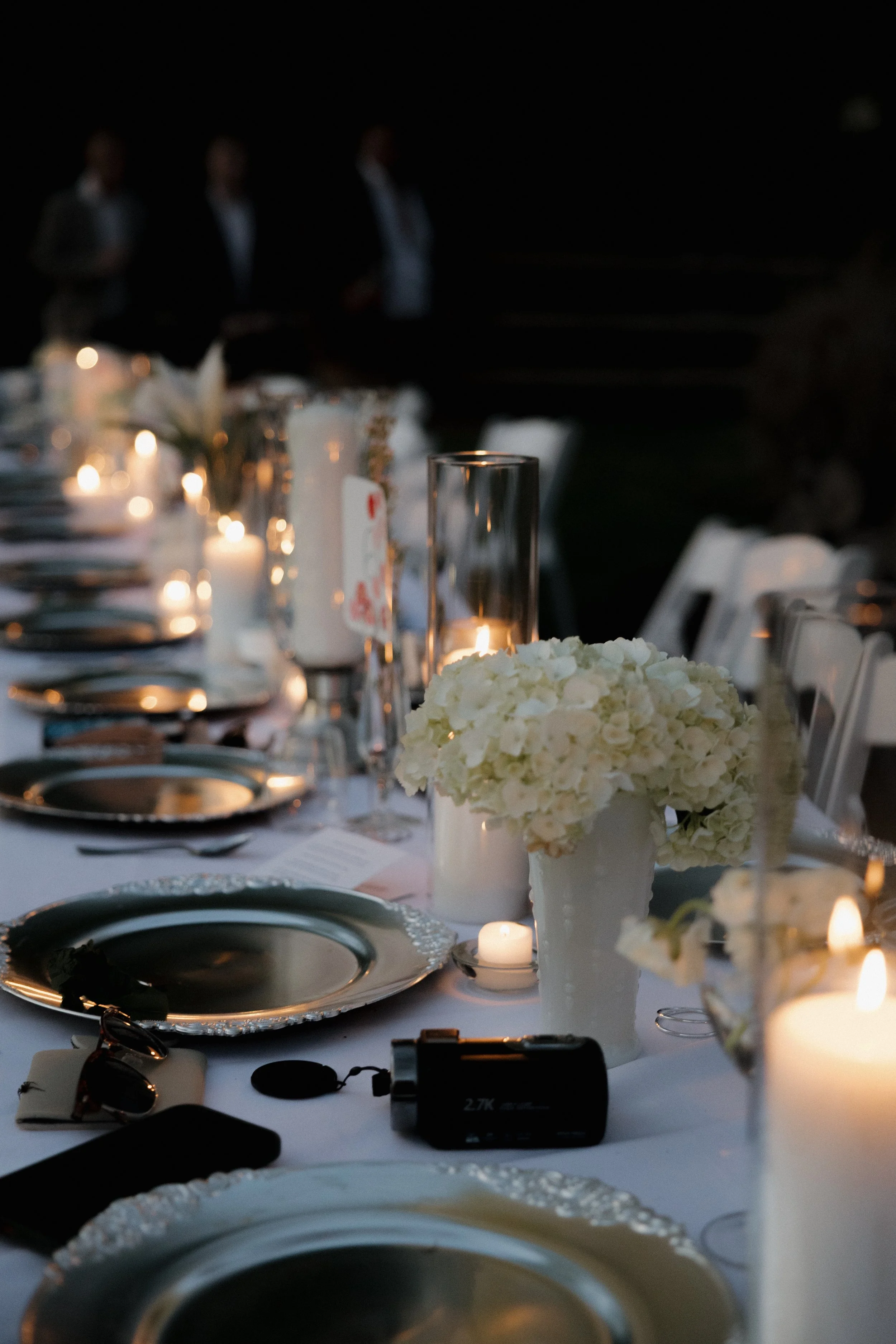 Elegant outdoor dining table set for a formal event at night, decorated with white flowers, candles, and silver plates, with blurred figures in the background.