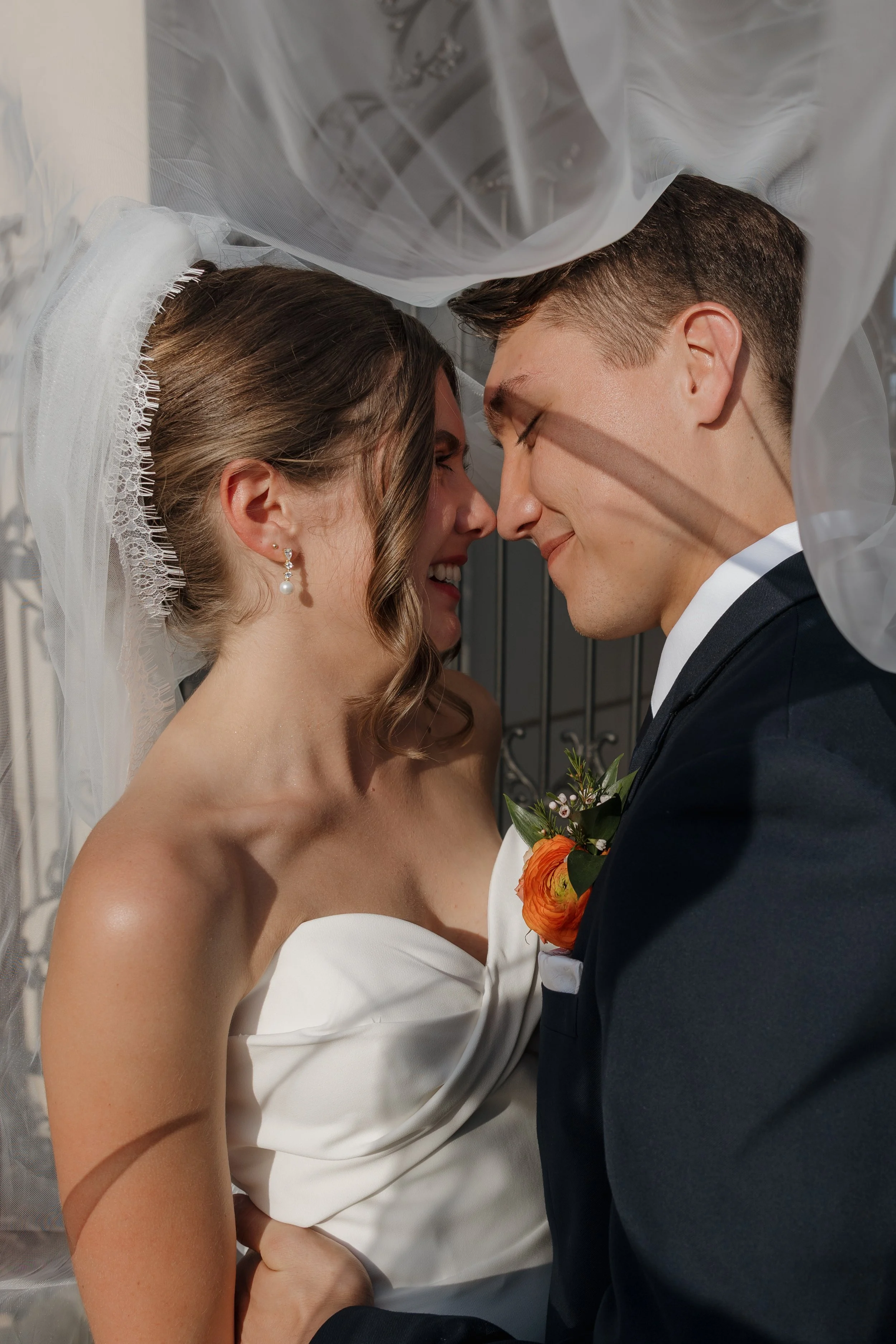 A bride and groom are smiling and leaning their foreheads together, captured in an intimate moment beneath the bride's veil on their wedding day.