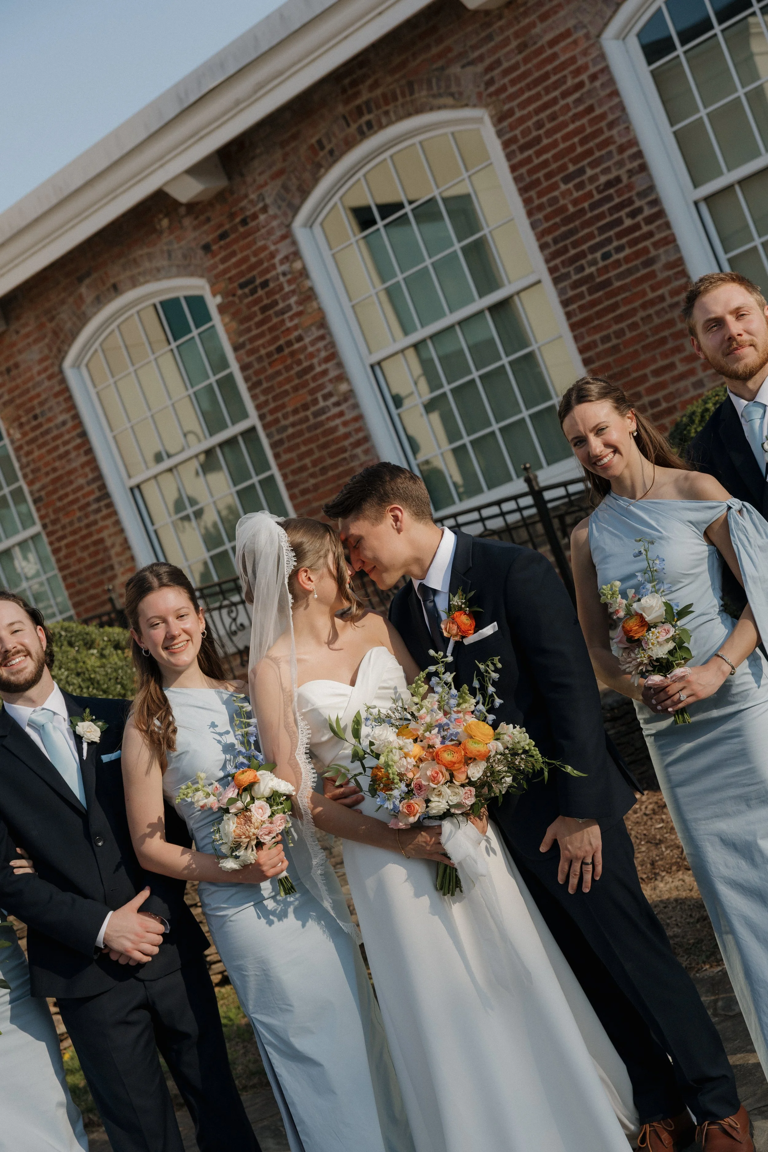 A wedding party with a bride, groom, and guests standing outside in front of a brick building with large windows, during daytime.