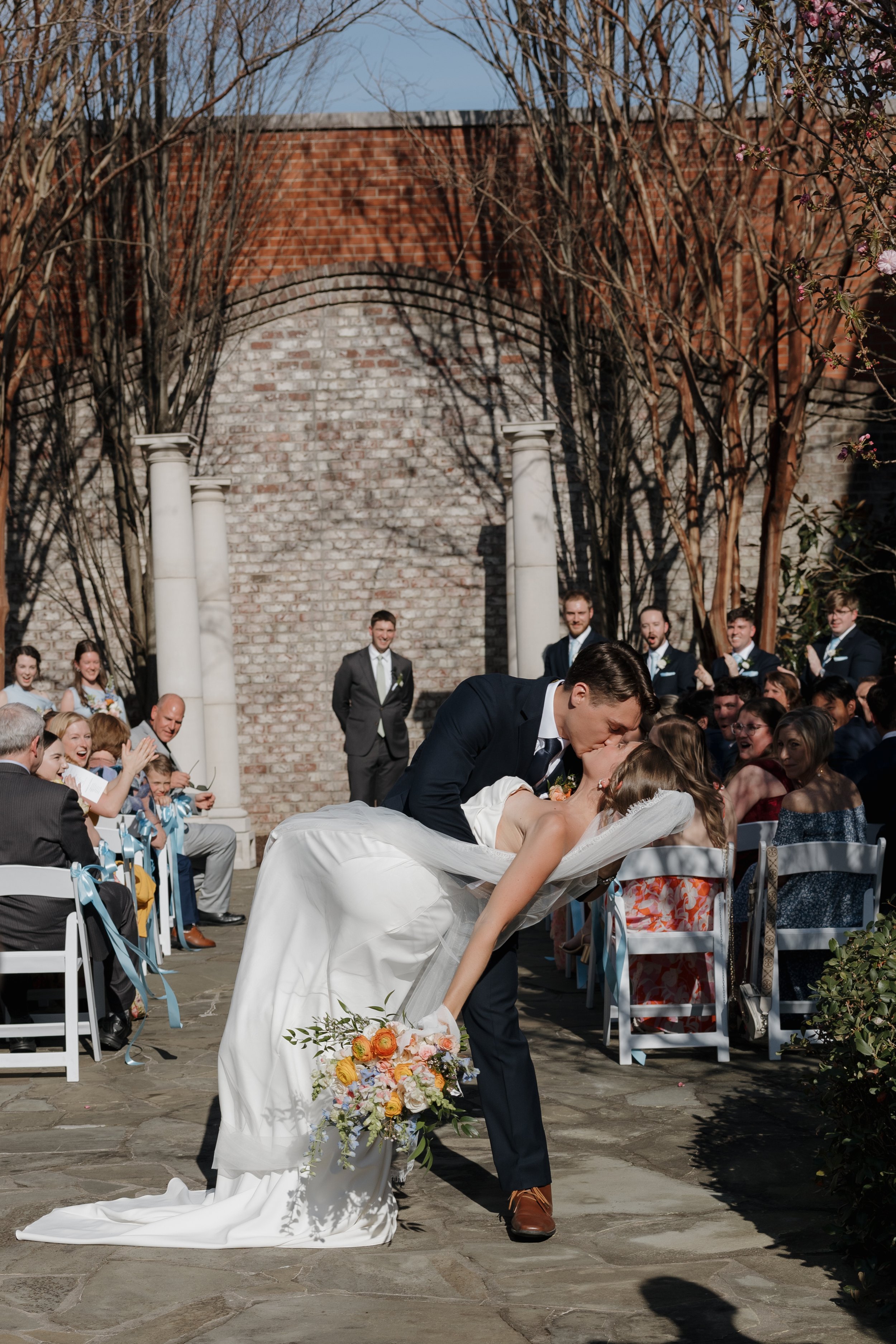 A couple sharing a kiss during their wedding ceremony outdoors, with guests seated on either side, a brick wall, white columns, and trees in the background.