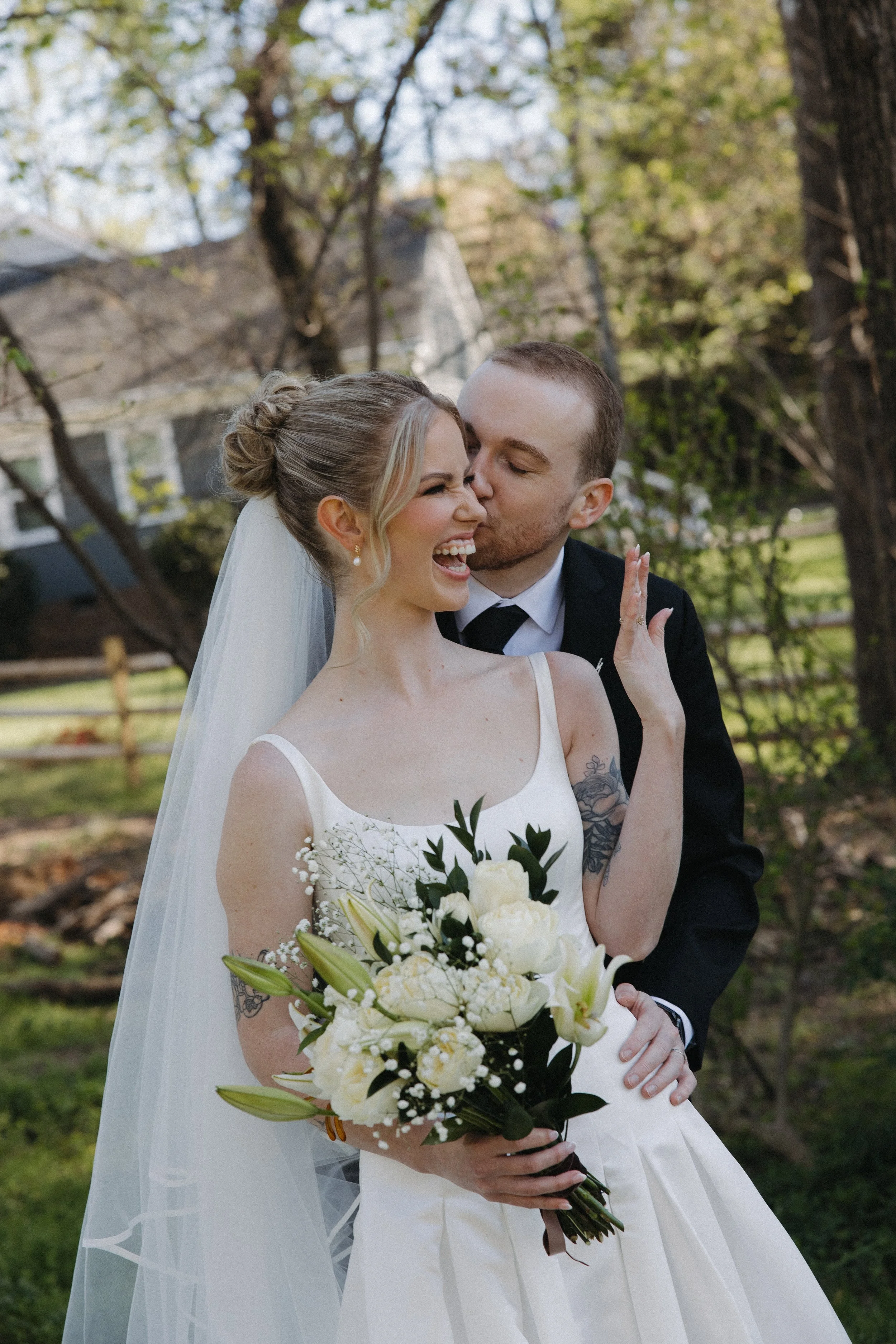 A newlywed couple celebrating outdoors, with the groom kissing the bride on her cheek. The bride is smiling and holding a bouquet of white flowers, with trees and a house in the background.