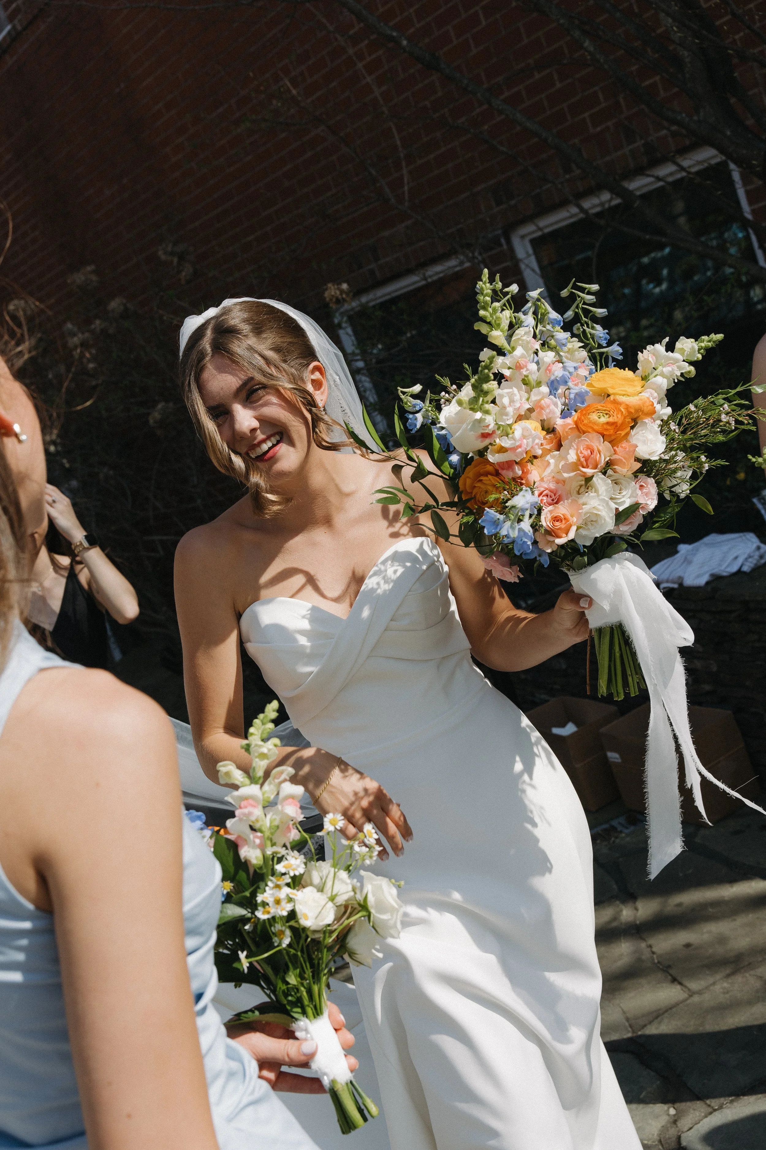 A bride with shoulder-length brown hair in a white wedding dress holds a large bouquet of colorful flowers, smiling happily during an outdoor wedding celebration.