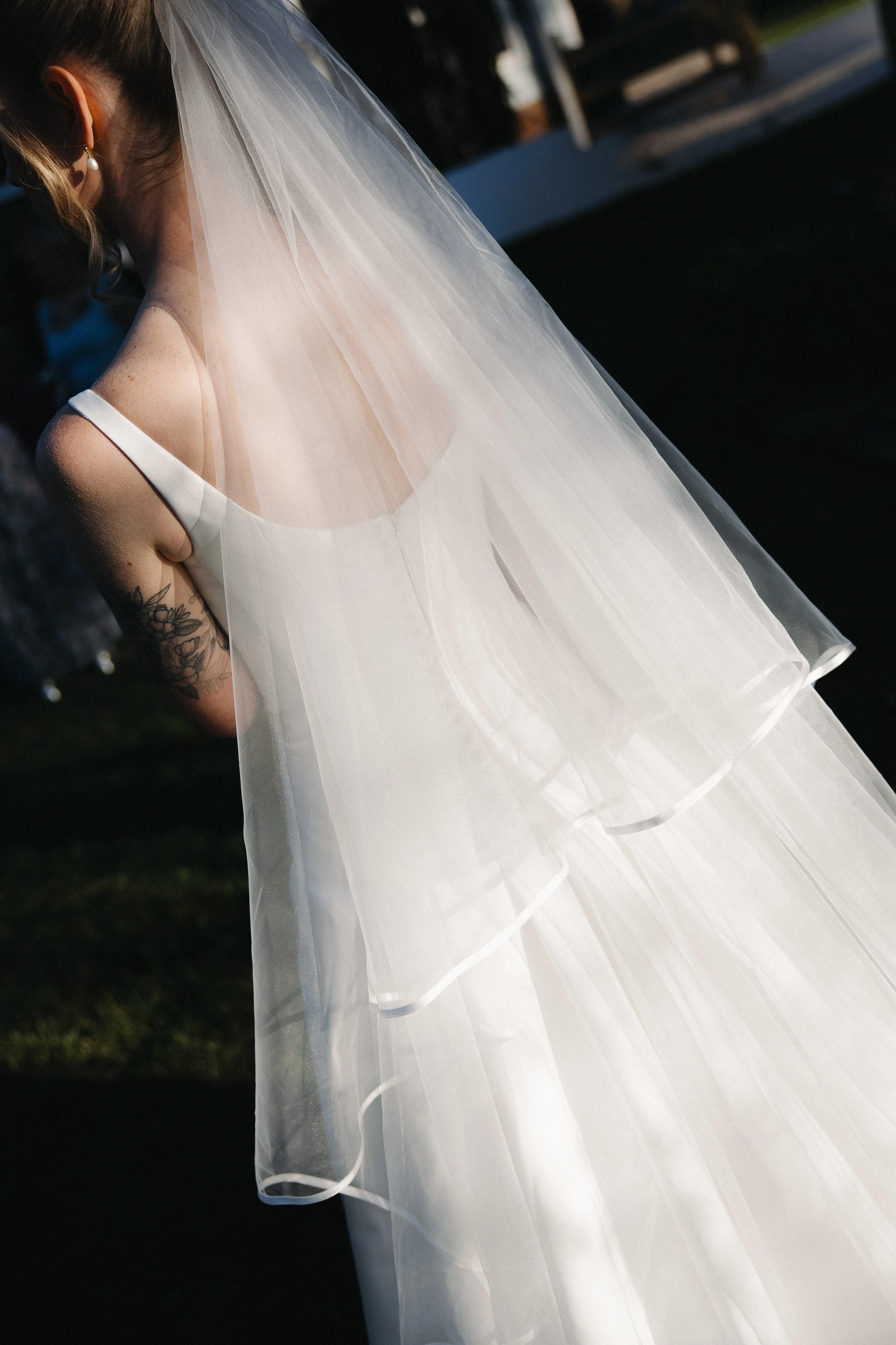 A bride wearing a white wedding dress with a sheer veil, showing a tattoo on her upper arm, outside during the day.