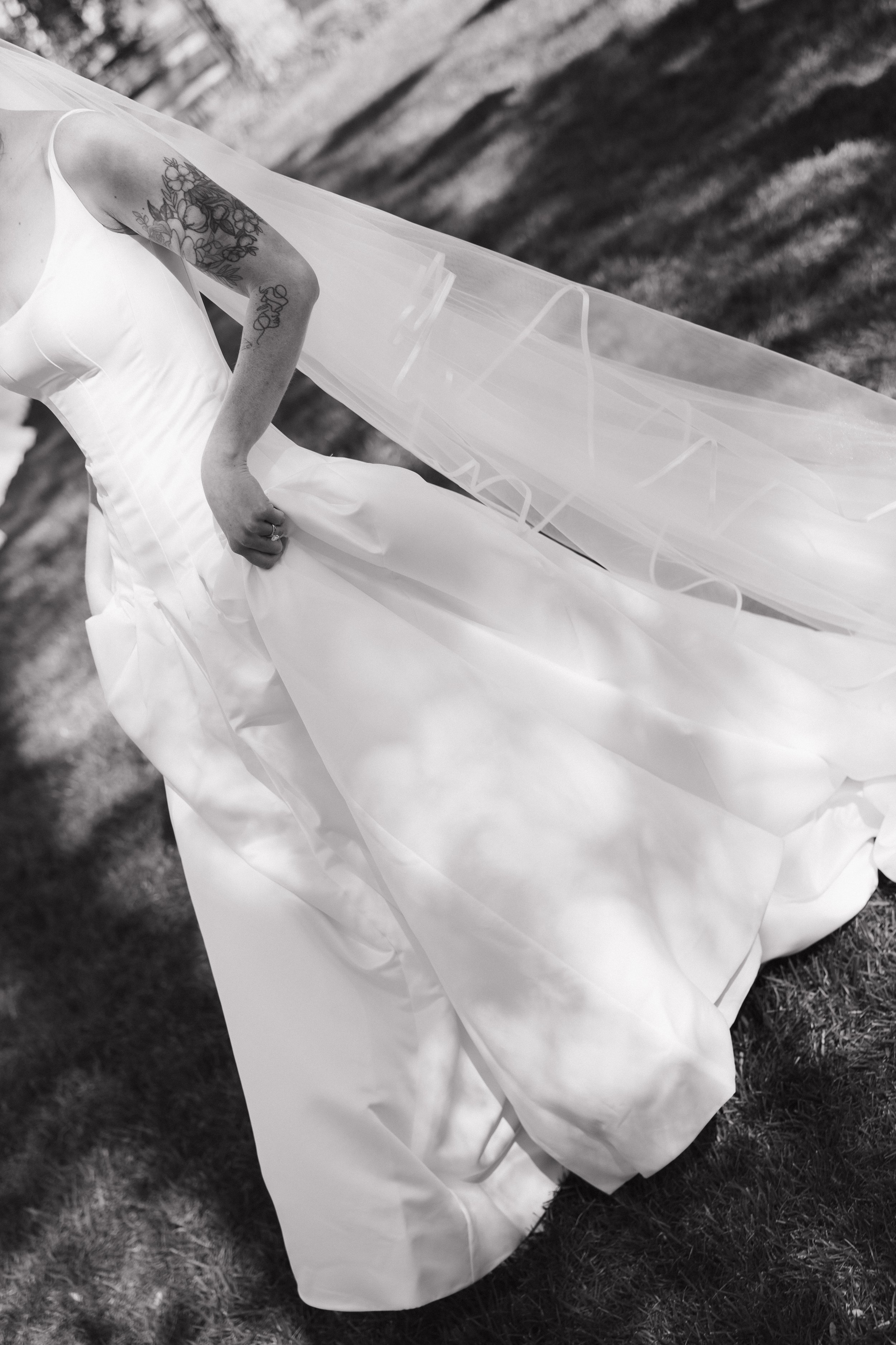 A person wearing a wedding dress with tattoos on their arm, standing outdoors on grass, holding the skirt of the dress.