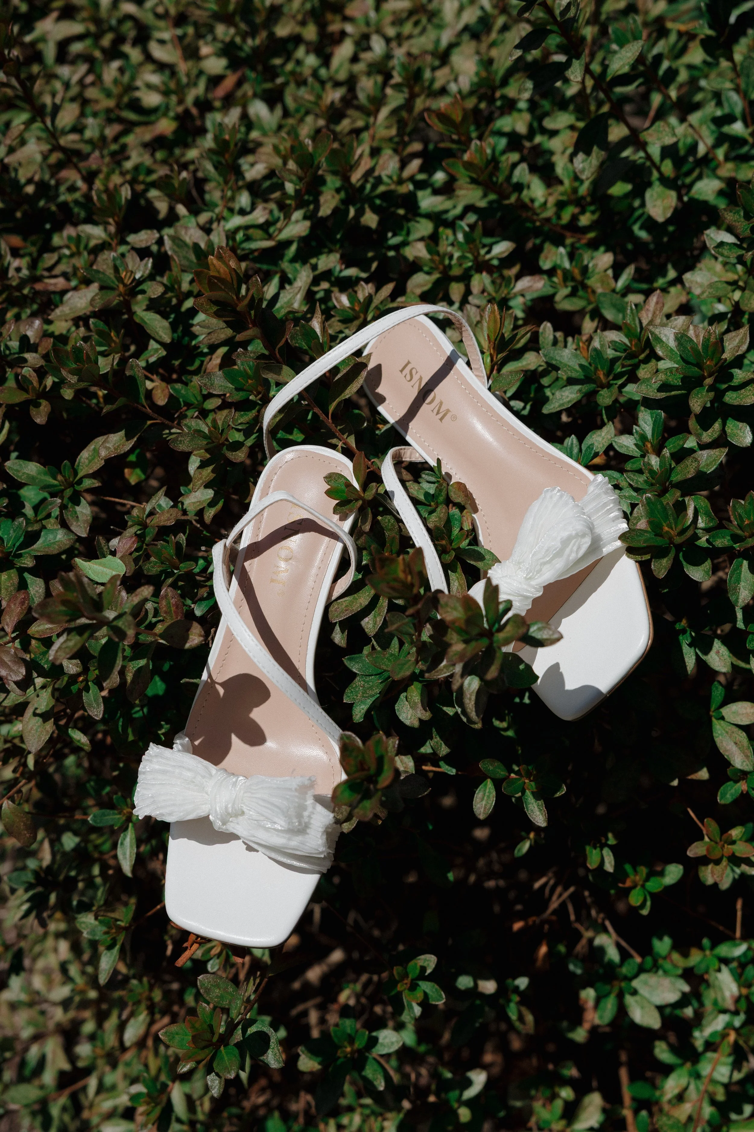 White high-heeled sandals with ankle straps and tied white bows, resting on green leafy bushes.