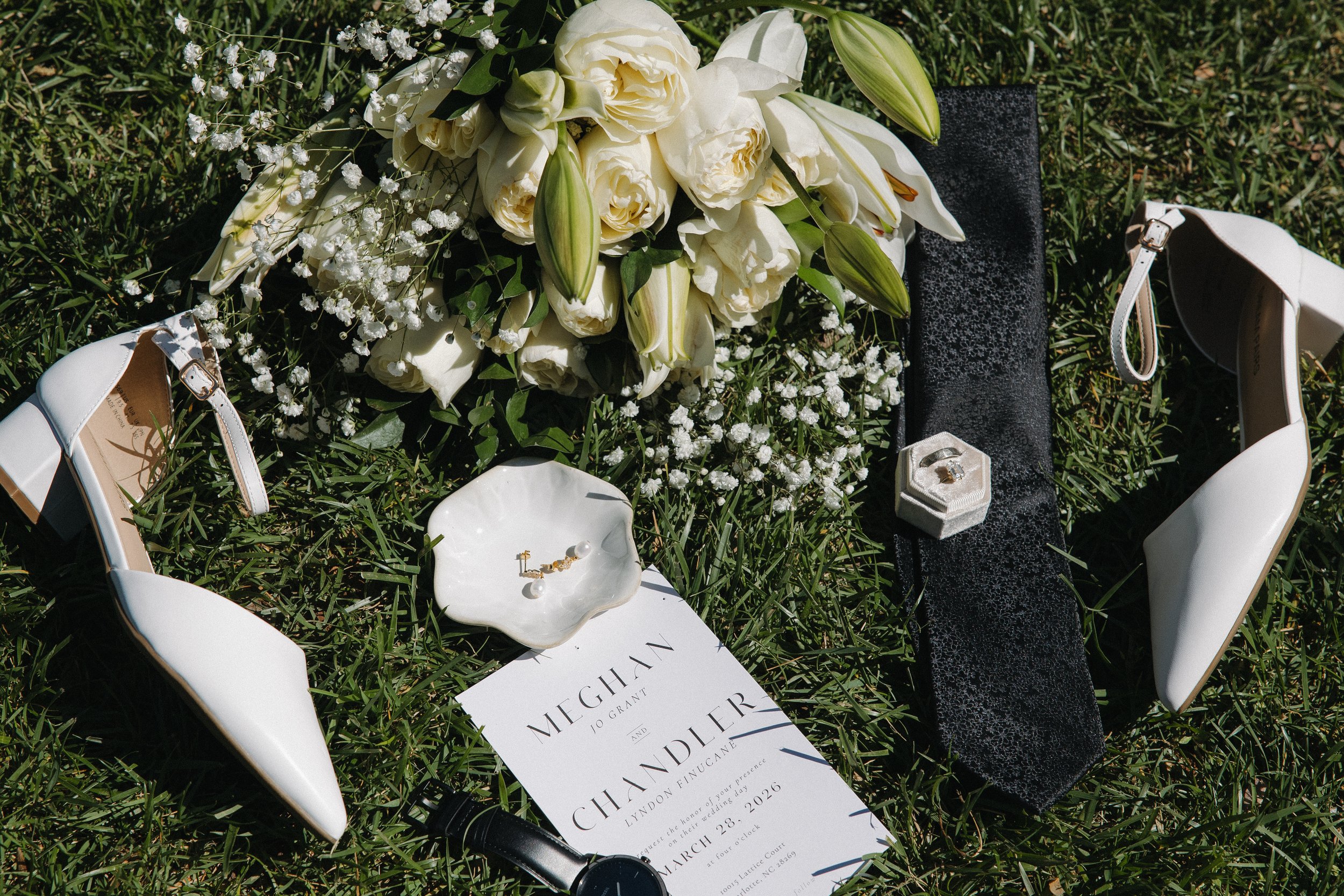Wedding bouquet with white flowers, white high-heeled shoes, a black fabric, a wedding invitation for Meghan and Chandler's wedding, a small white dish with jewelry, a black watch, and a ring box with a ring, all arranged on grass.