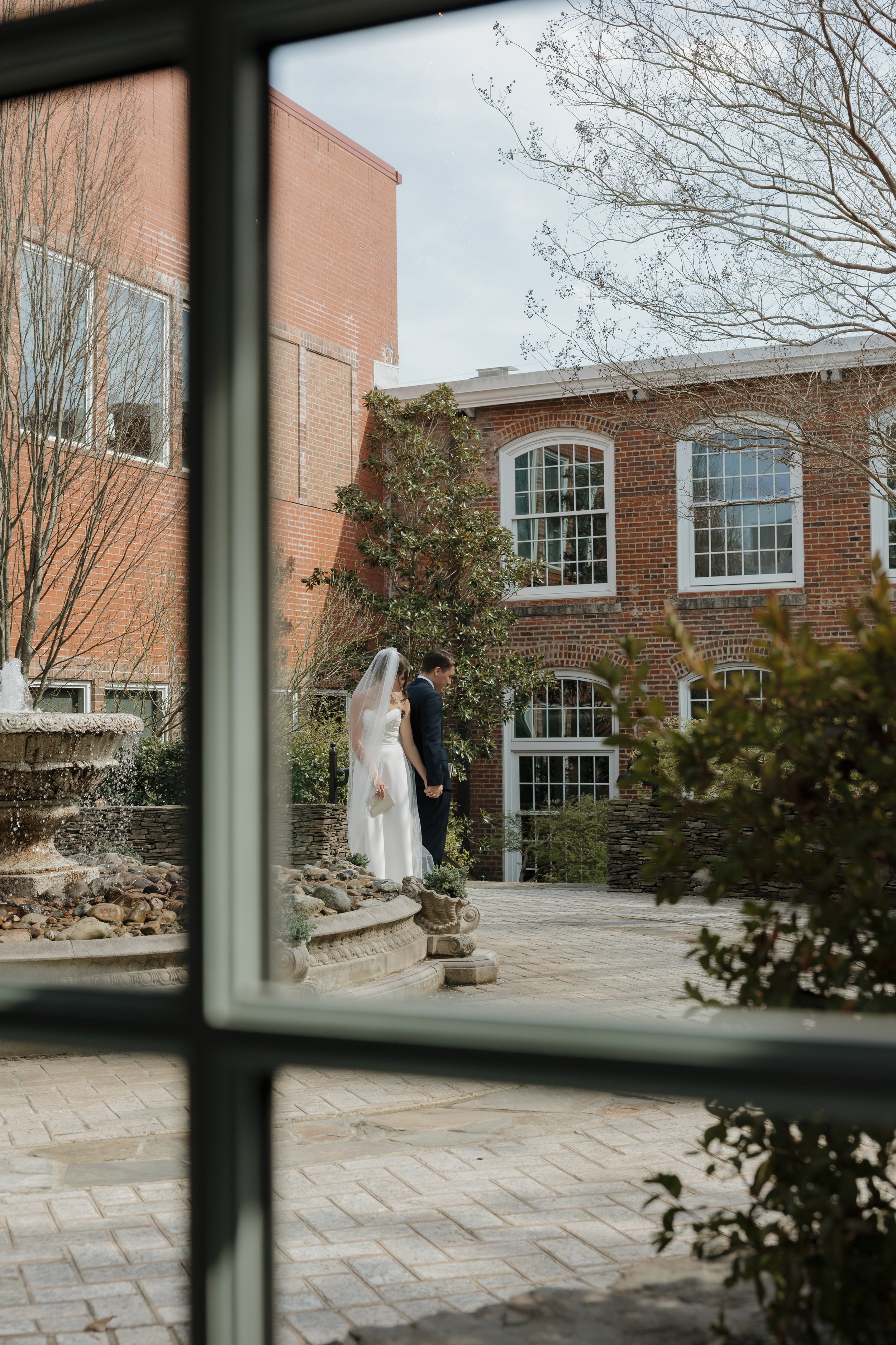 A bride and groom holding hands outdoors near a decorative fountain and brick building, viewed through a window.