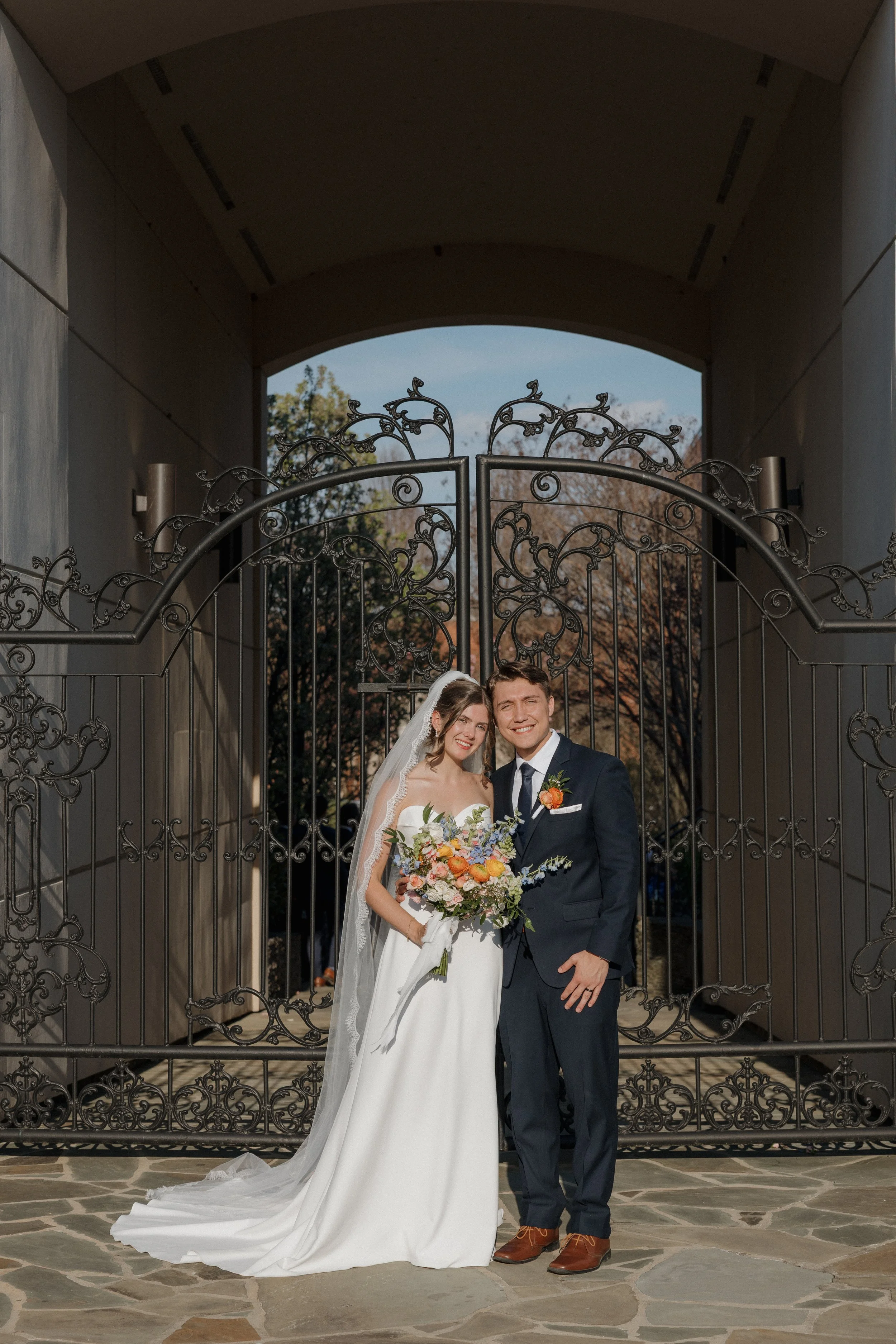 A bride and groom stand together outdoors in front of an ornate iron gate, smiling for a wedding photo. The bride wears a white wedding dress and veil, holding a colorful bouquet, while the groom wears a navy blue suit with a boutonniere.