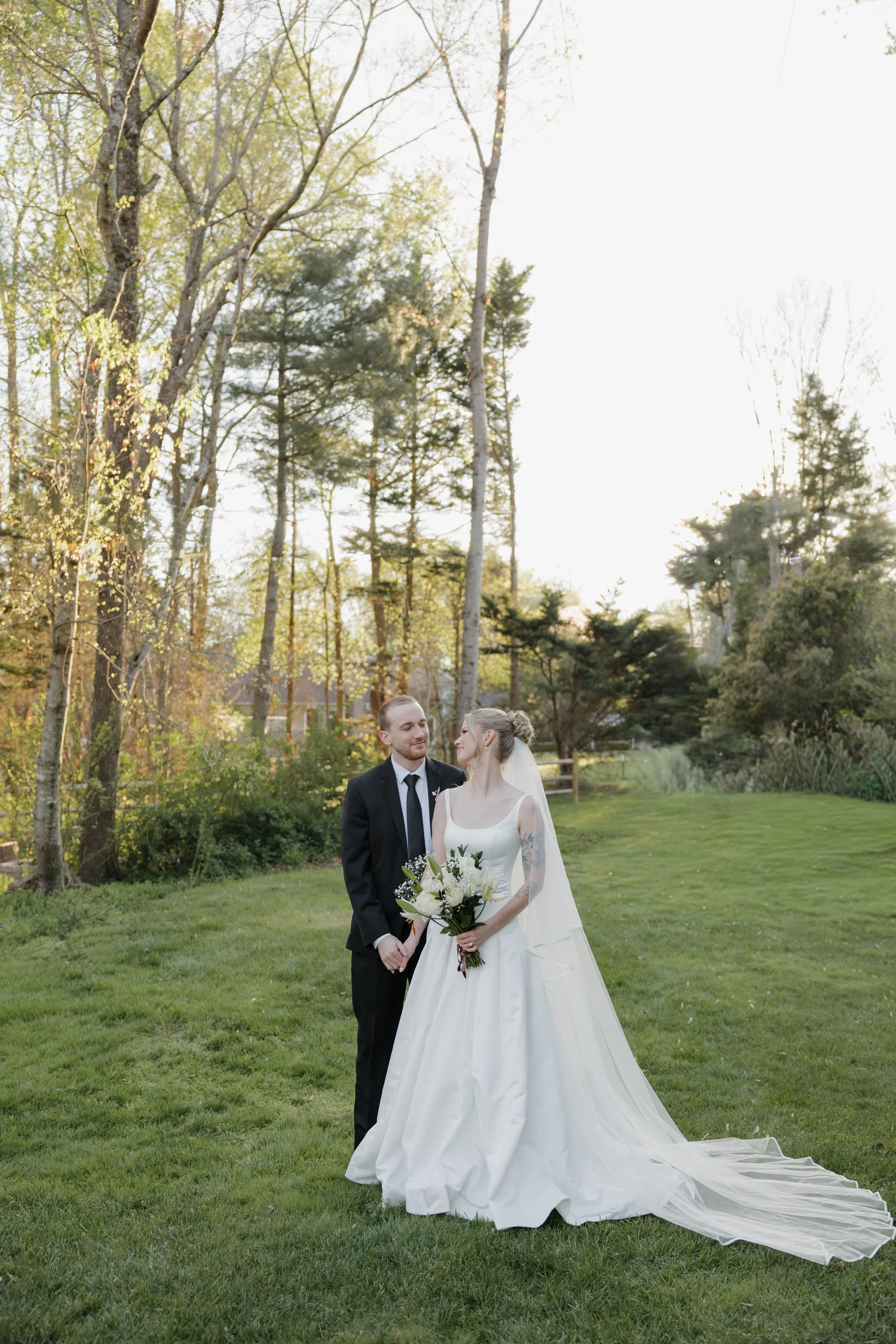 A bride and groom stand together outdoors in a grassy area surrounded by trees during sunset. The bride wears a white wedding gown with a long veil and holds a bouquet of white flowers. The groom wears a black suit with a white shirt and black tie, and they look at each other affectionately.
