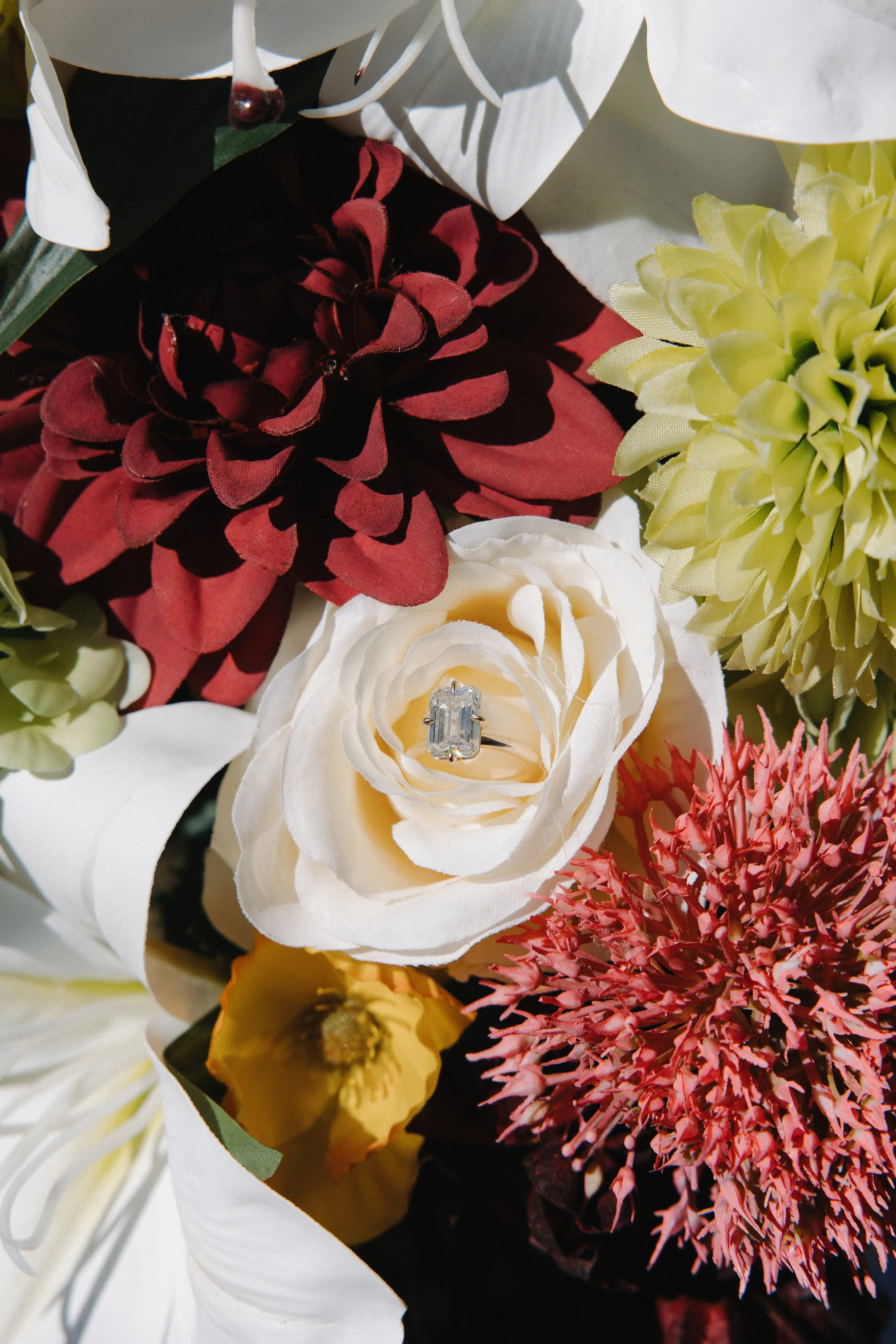 Close-up of a bouquet of artificial flowers, with a white rose featuring an engagement ring inside it, surrounded by red, yellow, and green flowers.