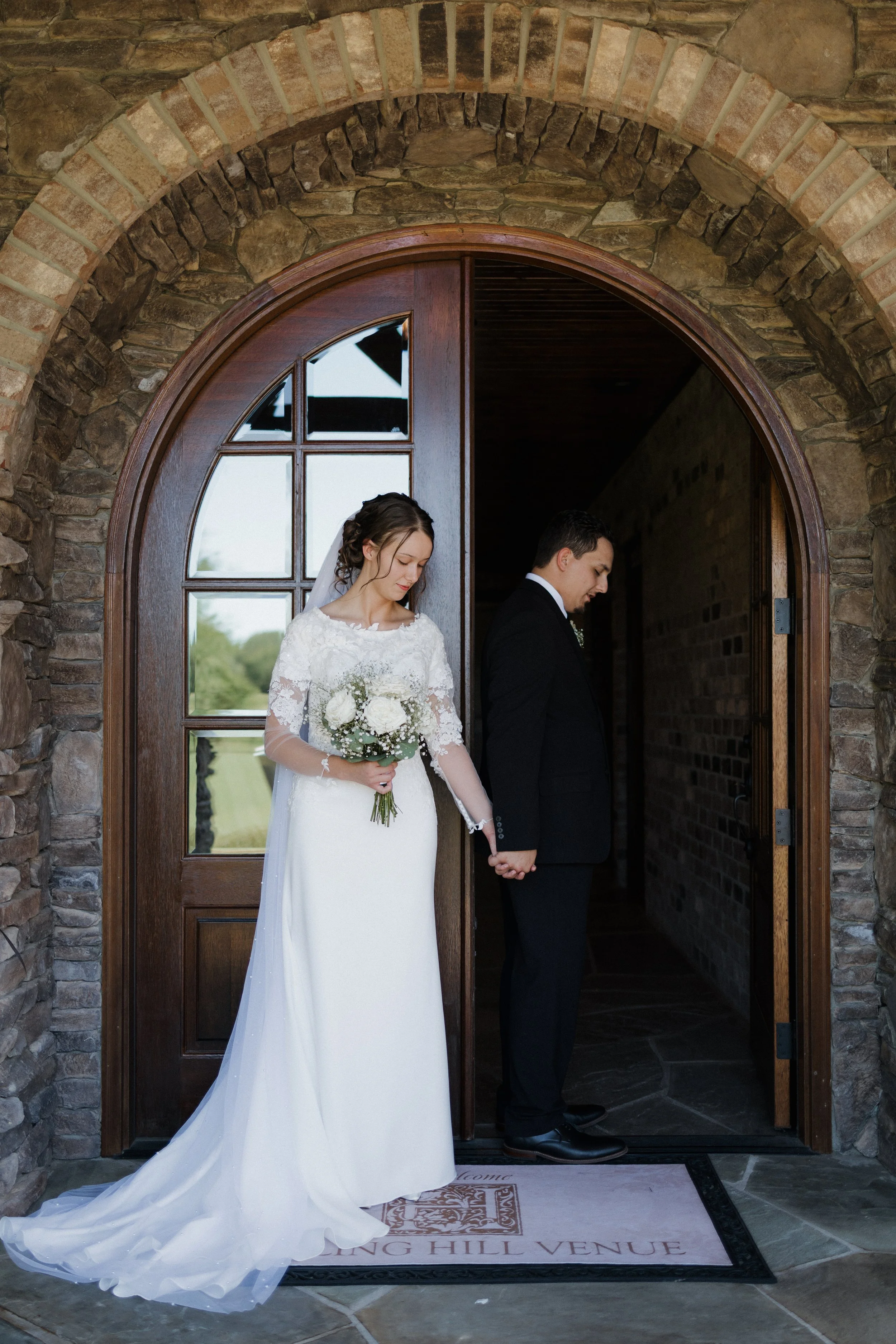 Bride and groom holding hands inside a stone and wood arched doorway, bride in a white lace wedding gown holding a bouquet, groom in a black tuxedo, standing on a welcome mat at a wedding venue.