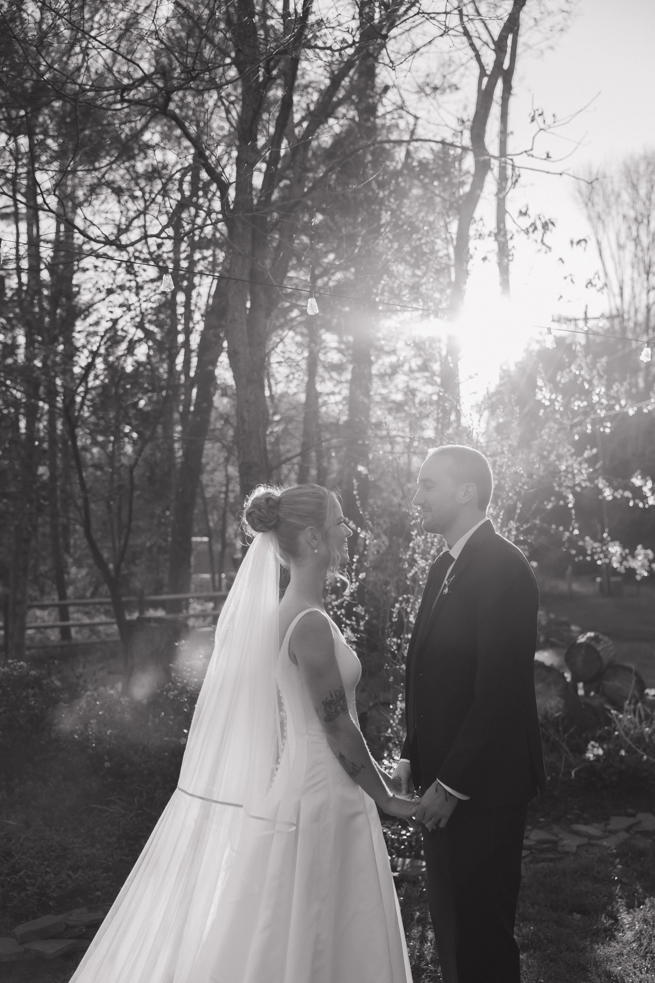 A couple dressed in wedding attire holding hands outdoors in a wooded area, with the sun setting behind them.
