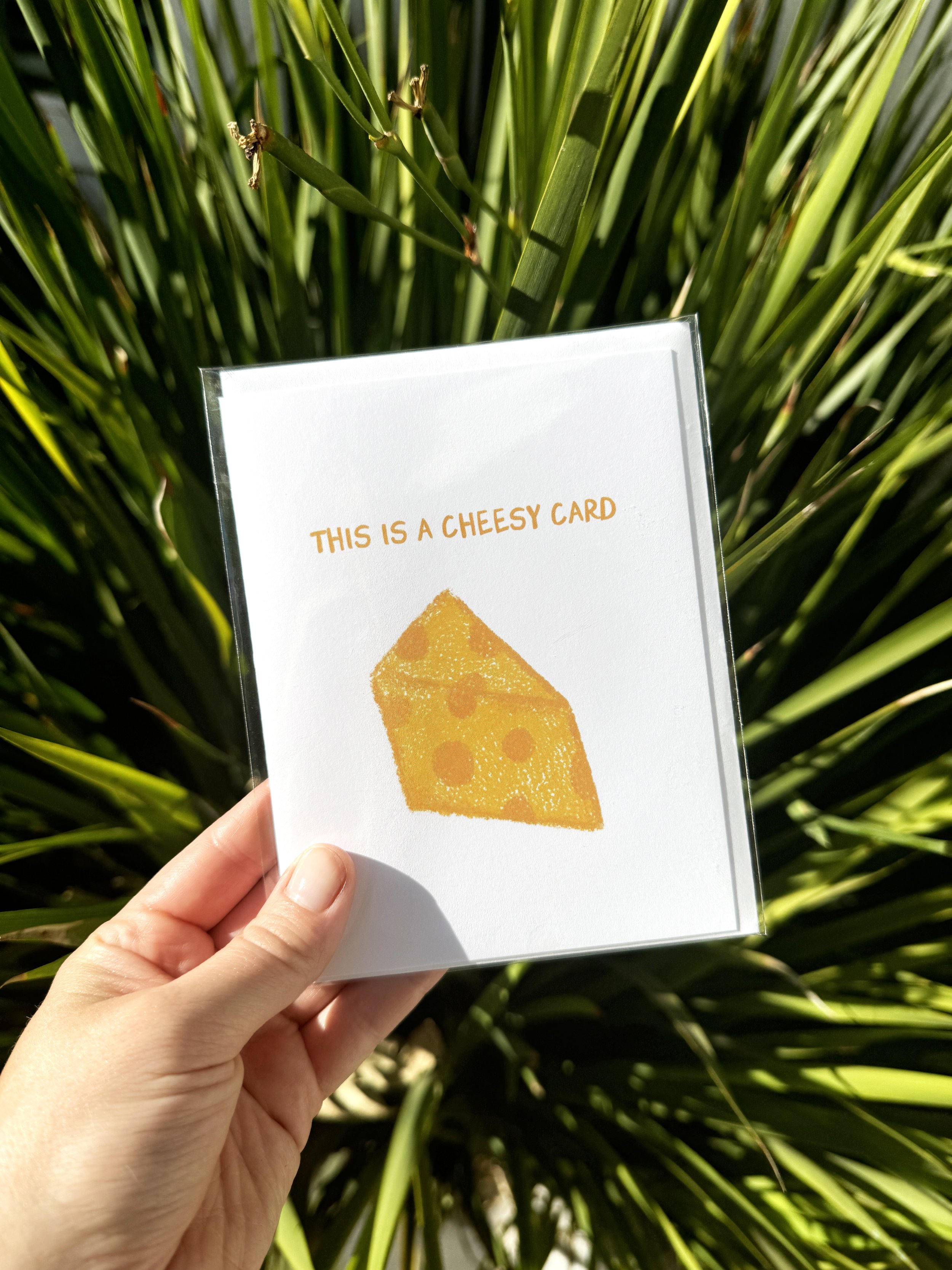 Hand holding greeting card that says, "this is a cheesy card" with an illustrated block of yellow cheese