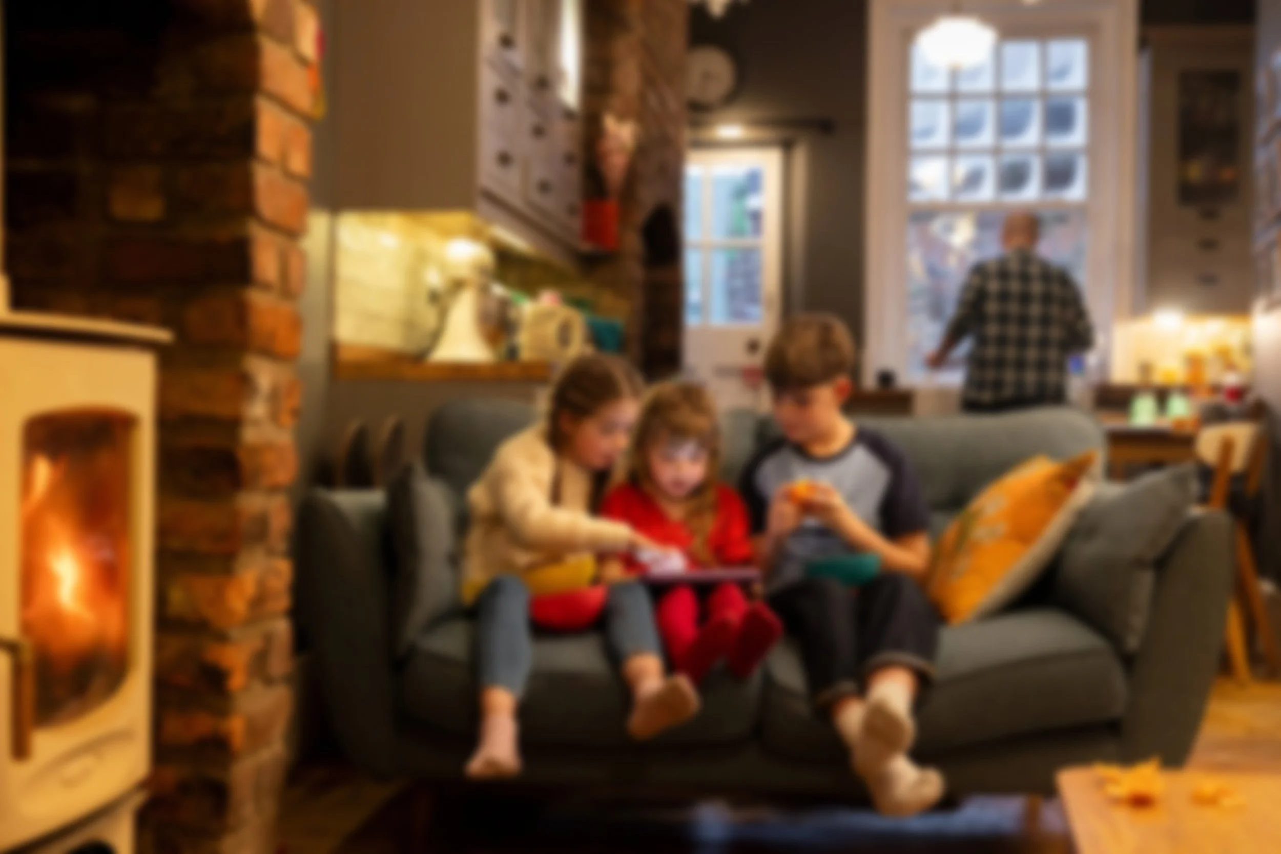Blurred photo of three children sitting on a gray couch, looking at a tablet and a phone inside a cozy living room with a fireplace, brick wall, and large windows.