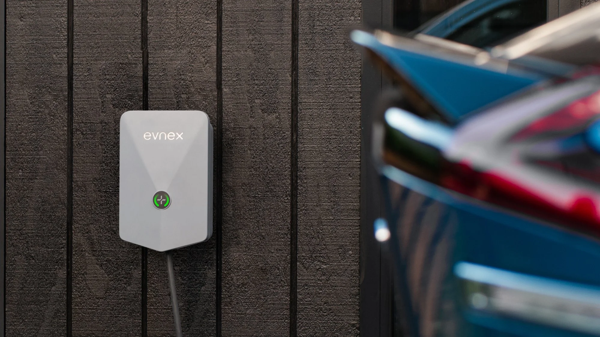 Electric vehicle charging station with a gray charger mounted on a dark wooden wall, and a car parked nearby.