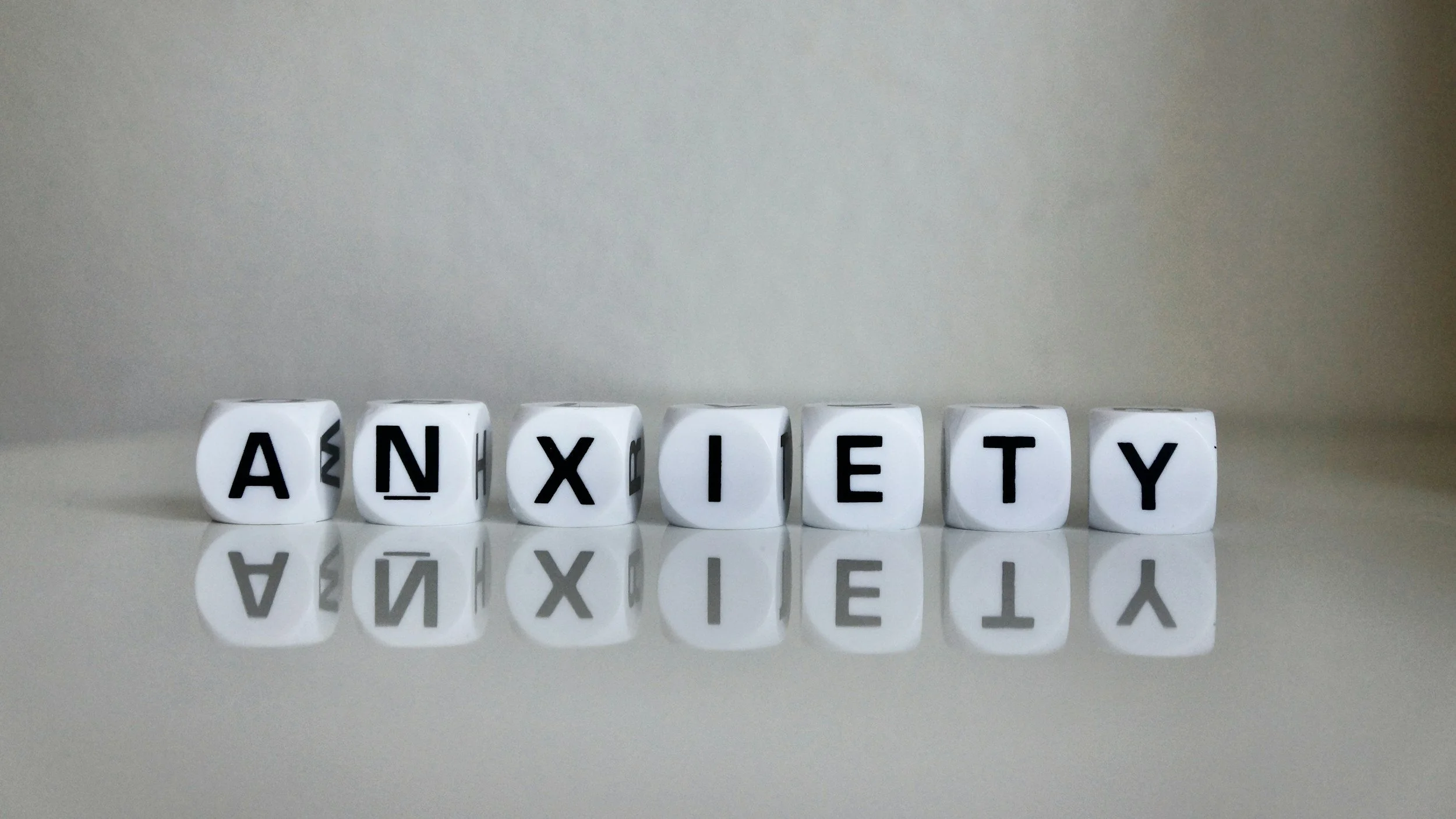 White dice with black letters spelling out 'ANXIETY' on a reflective surface against a plain light gray background.