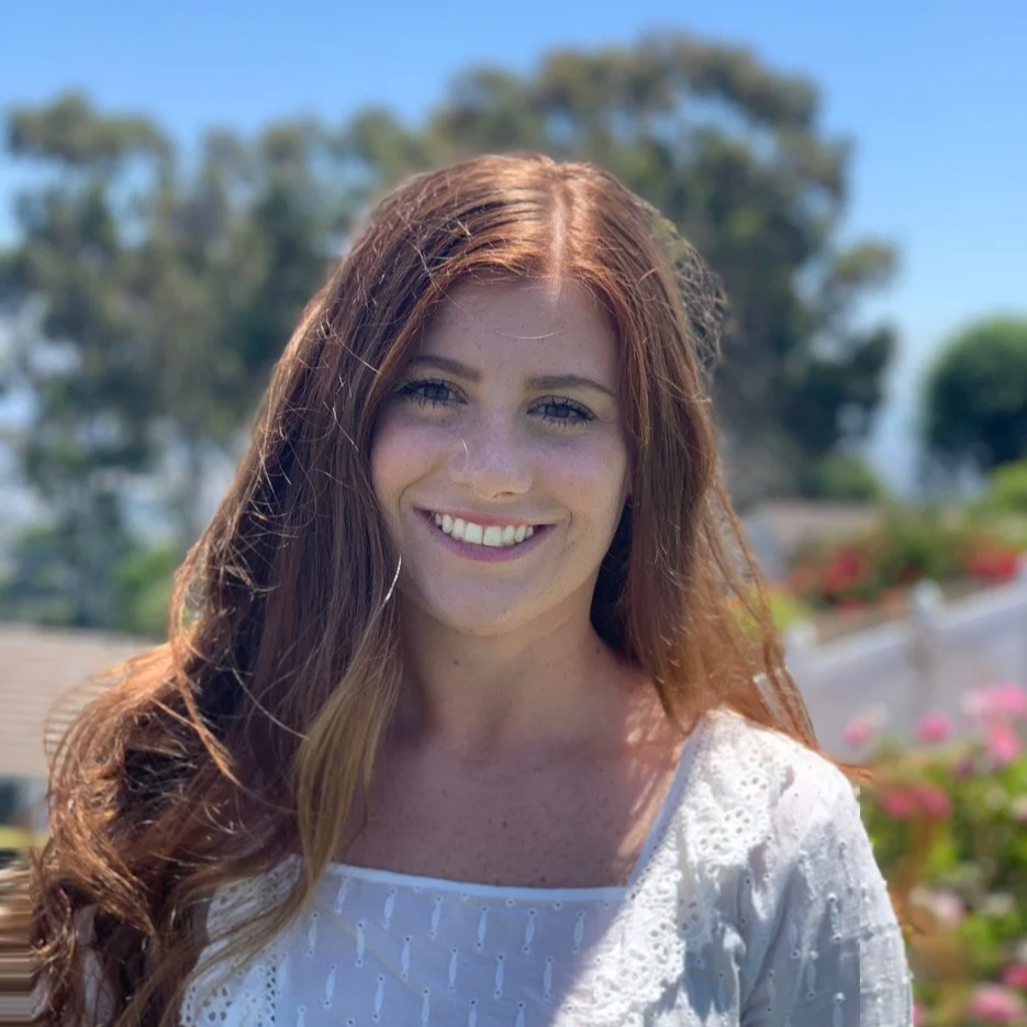 A young woman with long, wavy red hair smiling outdoors on a sunny day, with trees and pink flowers in the background.