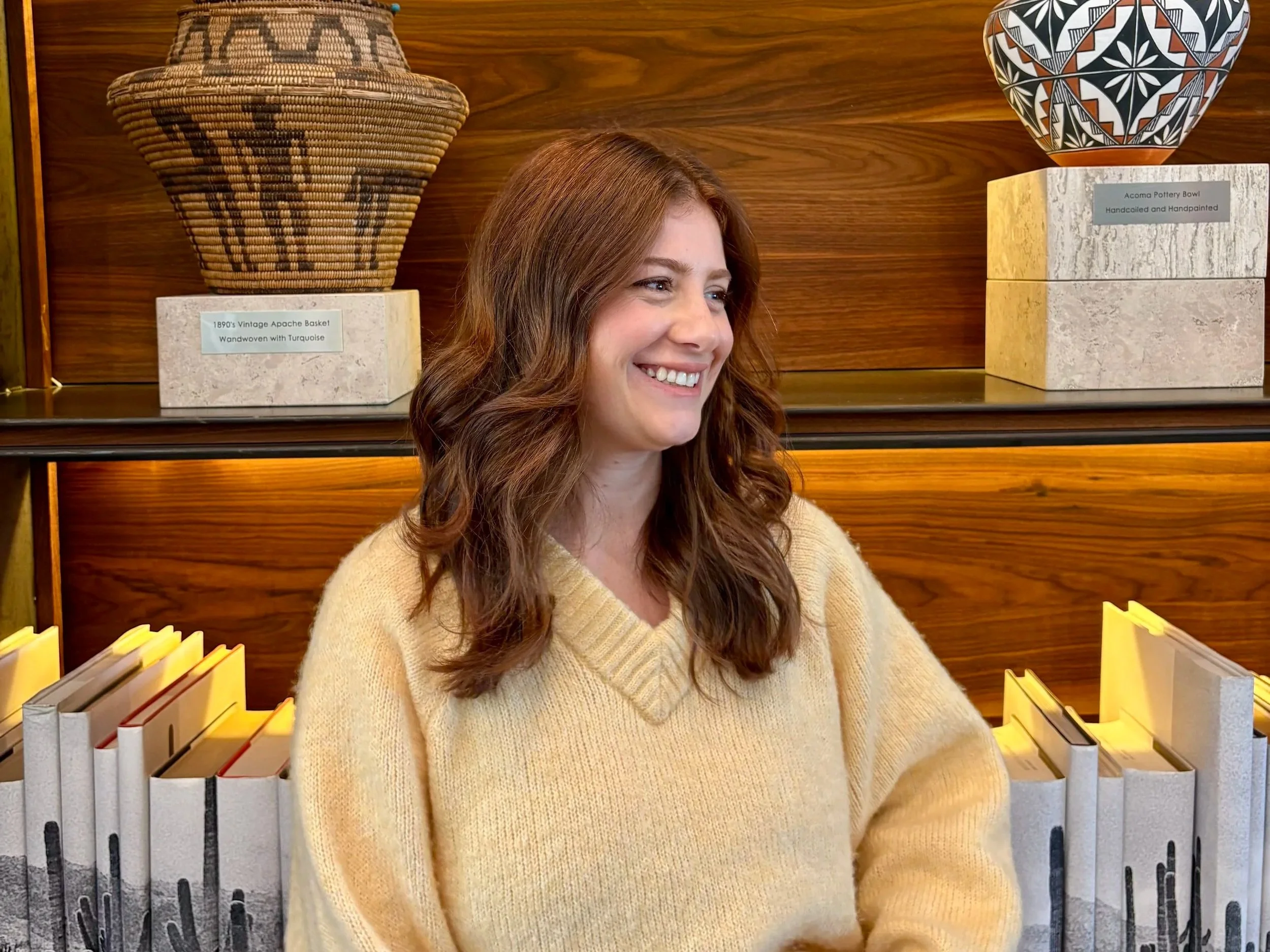 A woman with long brown wavy hair smiling, sitting in front of a wooden shelf with ceramic and woven baskets, wearing a cream-colored sweater.