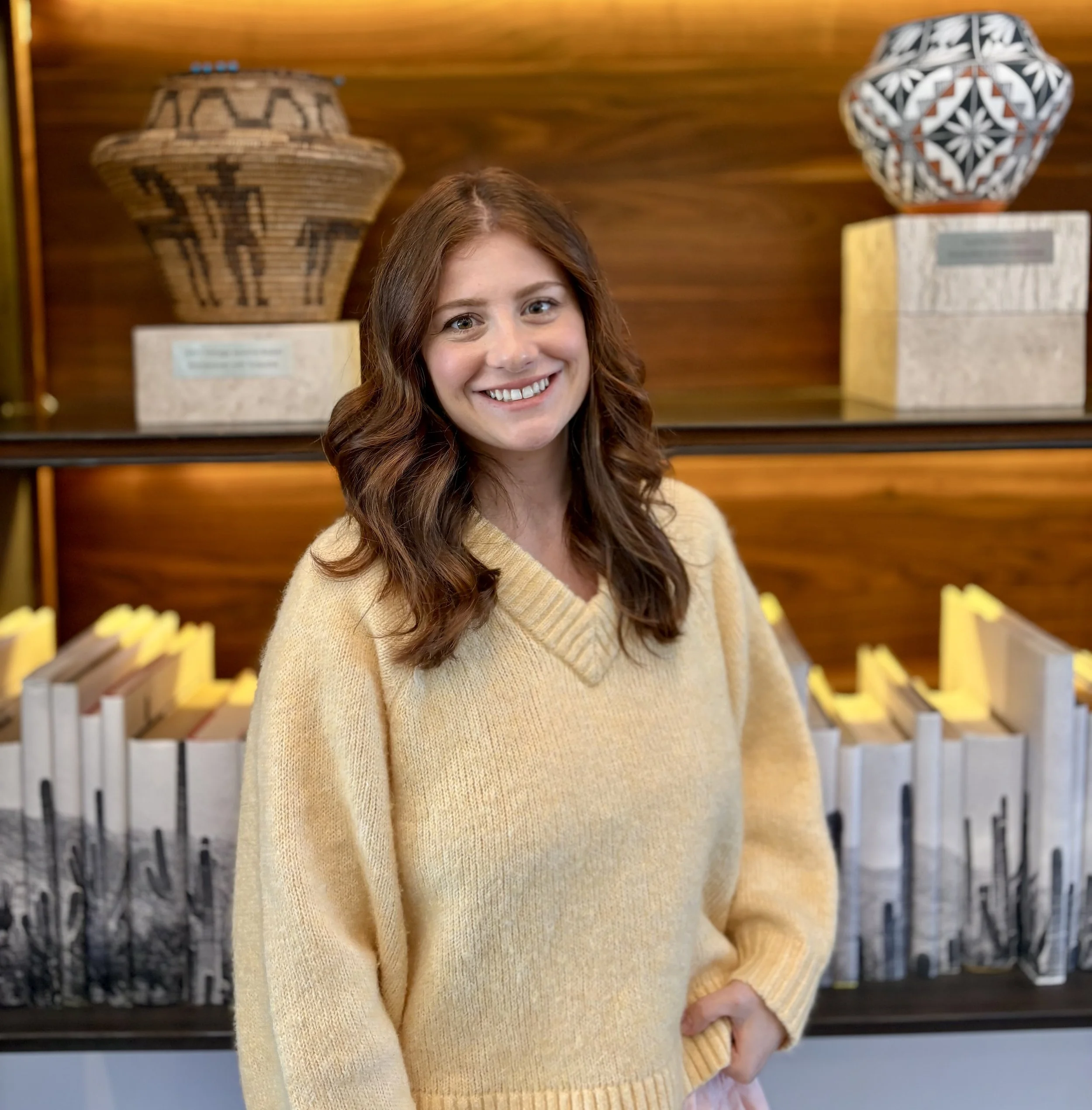 A smiling woman with wavy brown hair wearing a yellow sweater, standing in front of decorative bowls on a bookshelf.