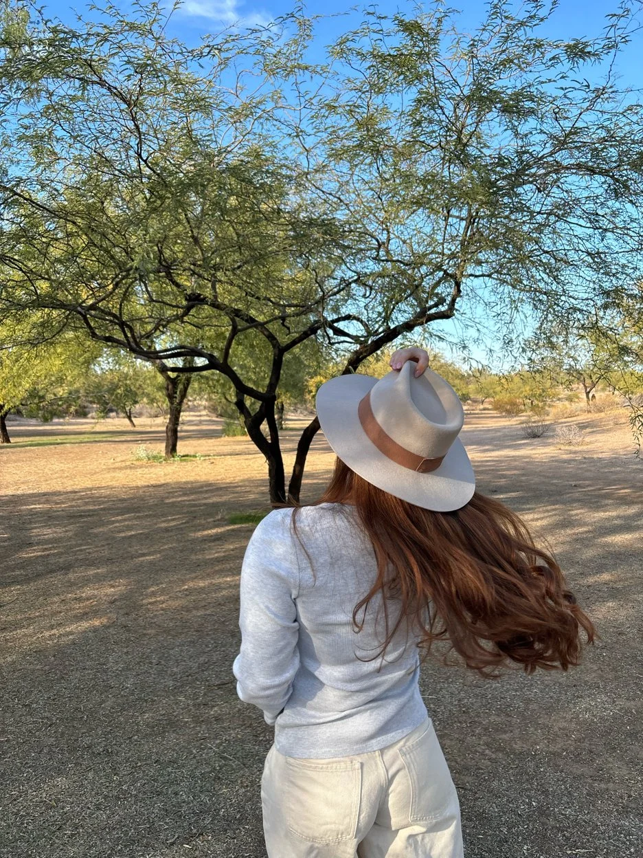 Woman with long red hair, wearing a gray sweater, cream-colored pants, and a beige sunhat, standing outdoors in a dry, desert-like landscape with sparse trees and a clear blue sky.
