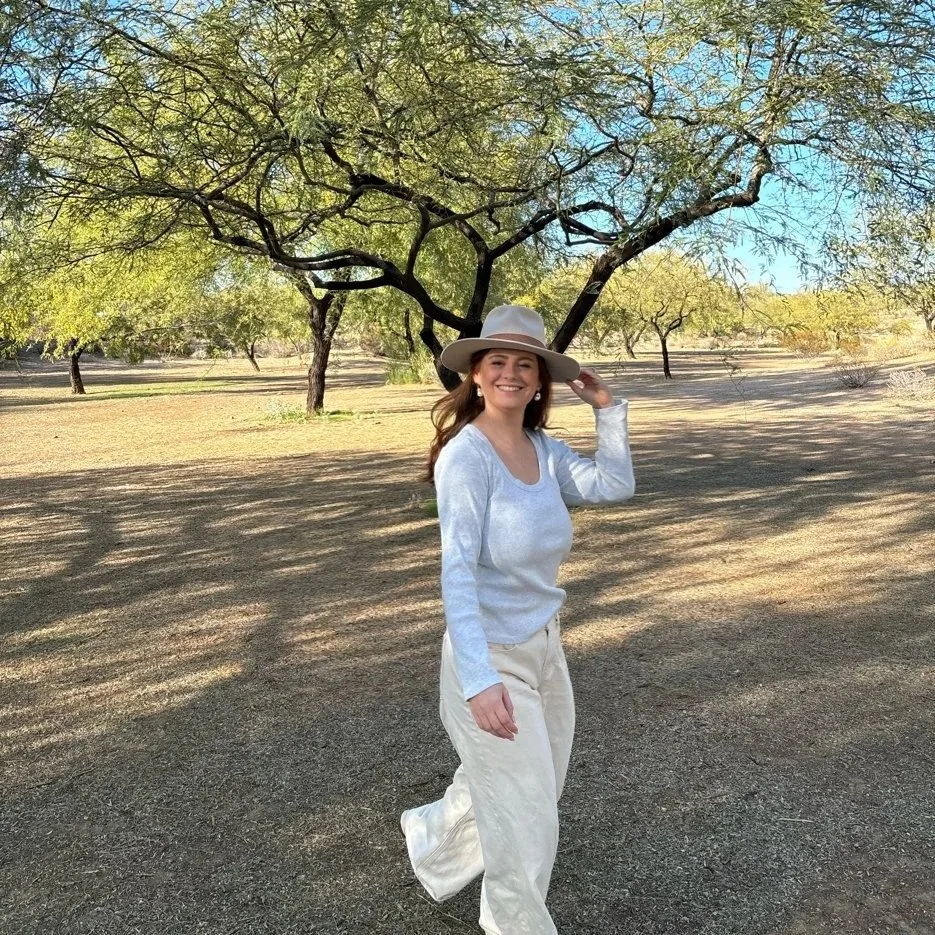 A woman wearing a white hat, white sweater, and cream-colored pants walking in a park with trees and gravel ground, smiling at the camera.