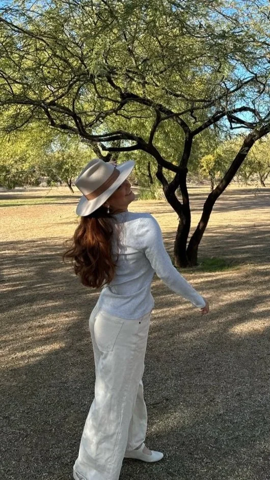A woman with long red hair wearing a white hat, white sweater, and white pants standing outdoors near a tree with green leaves in a sunny park.