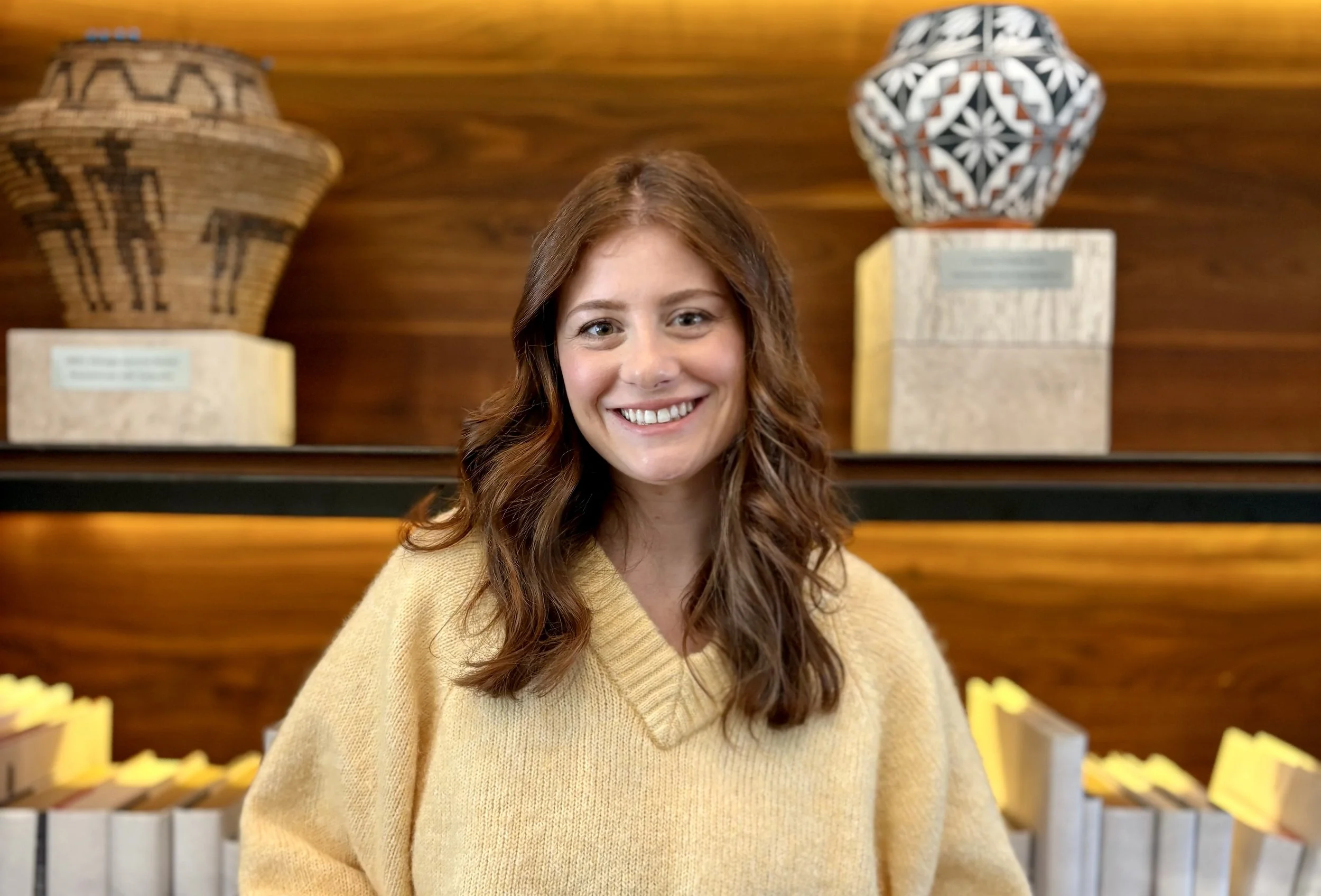 A smiling woman with wavy brown hair wearing a yellow sweater, standing in front of decorative pottery on display in a warm-toned room.