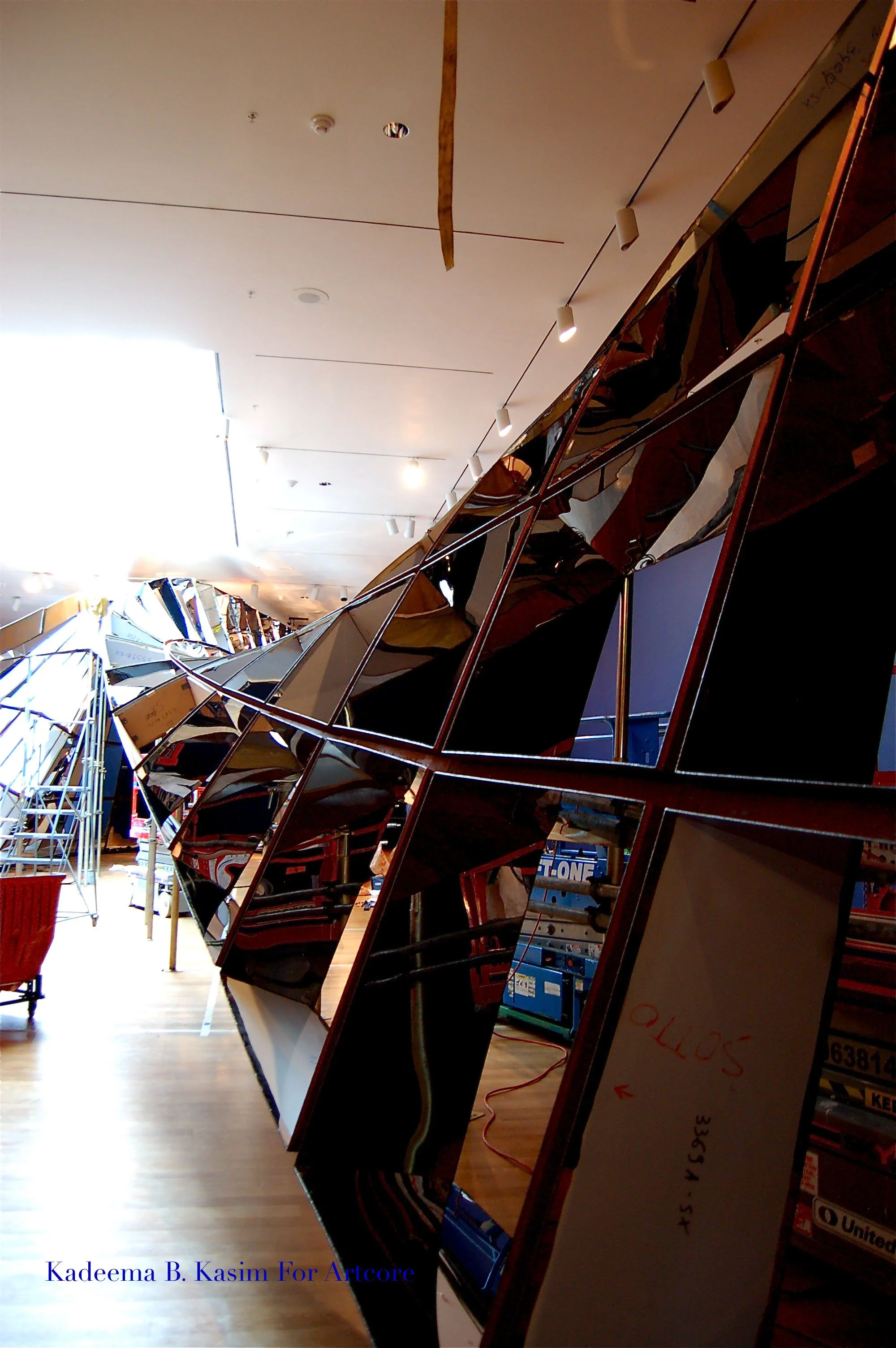 Art installation featuring a large, angled mirror with multiple reflective surfaces, creating a fragmented reflection of the surrounding space. The setting appears to be indoors, with an illuminated ceiling and other art pieces in the background.