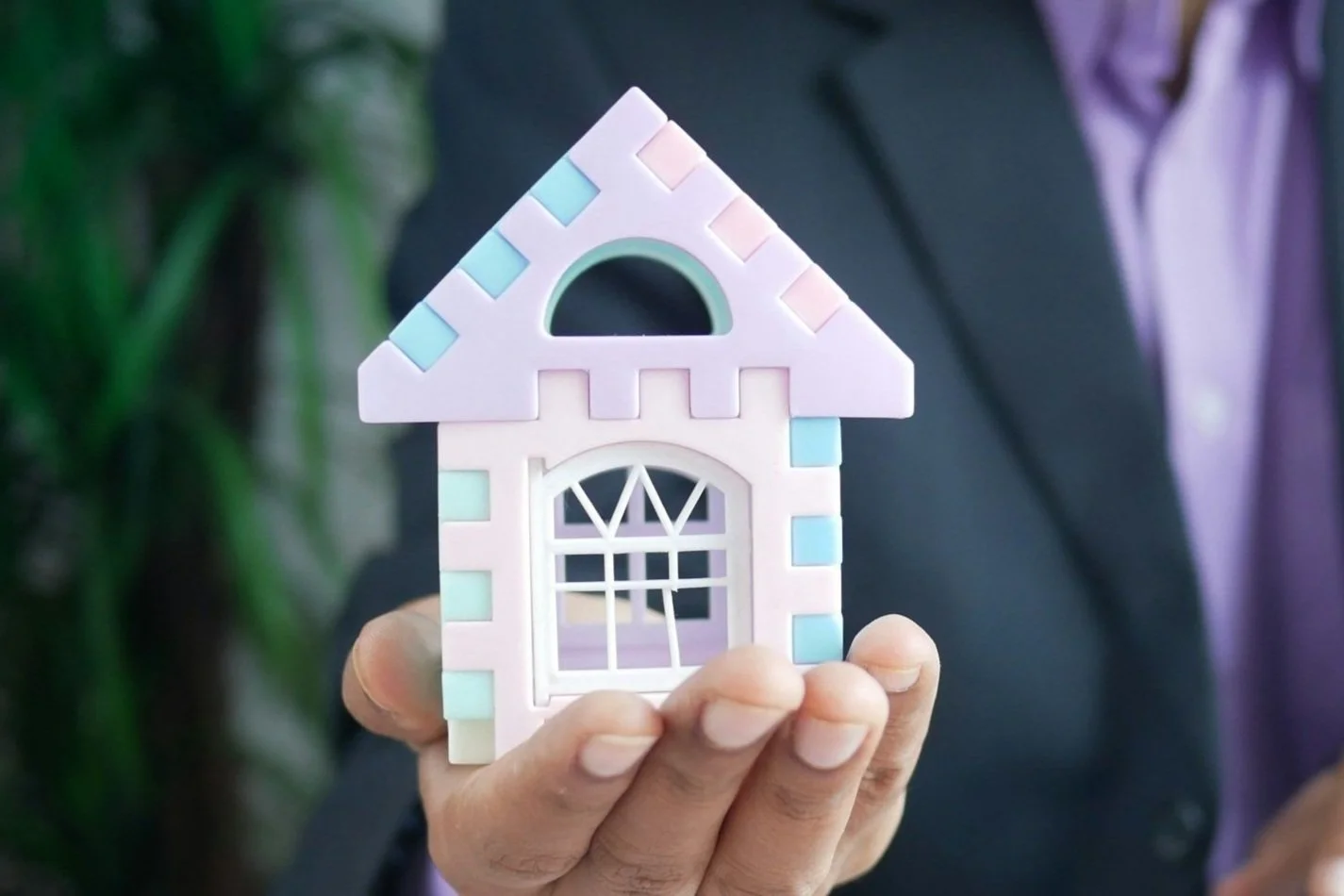 A person in business attire holding a colorful, miniature model of a house with a pink roof and window details.