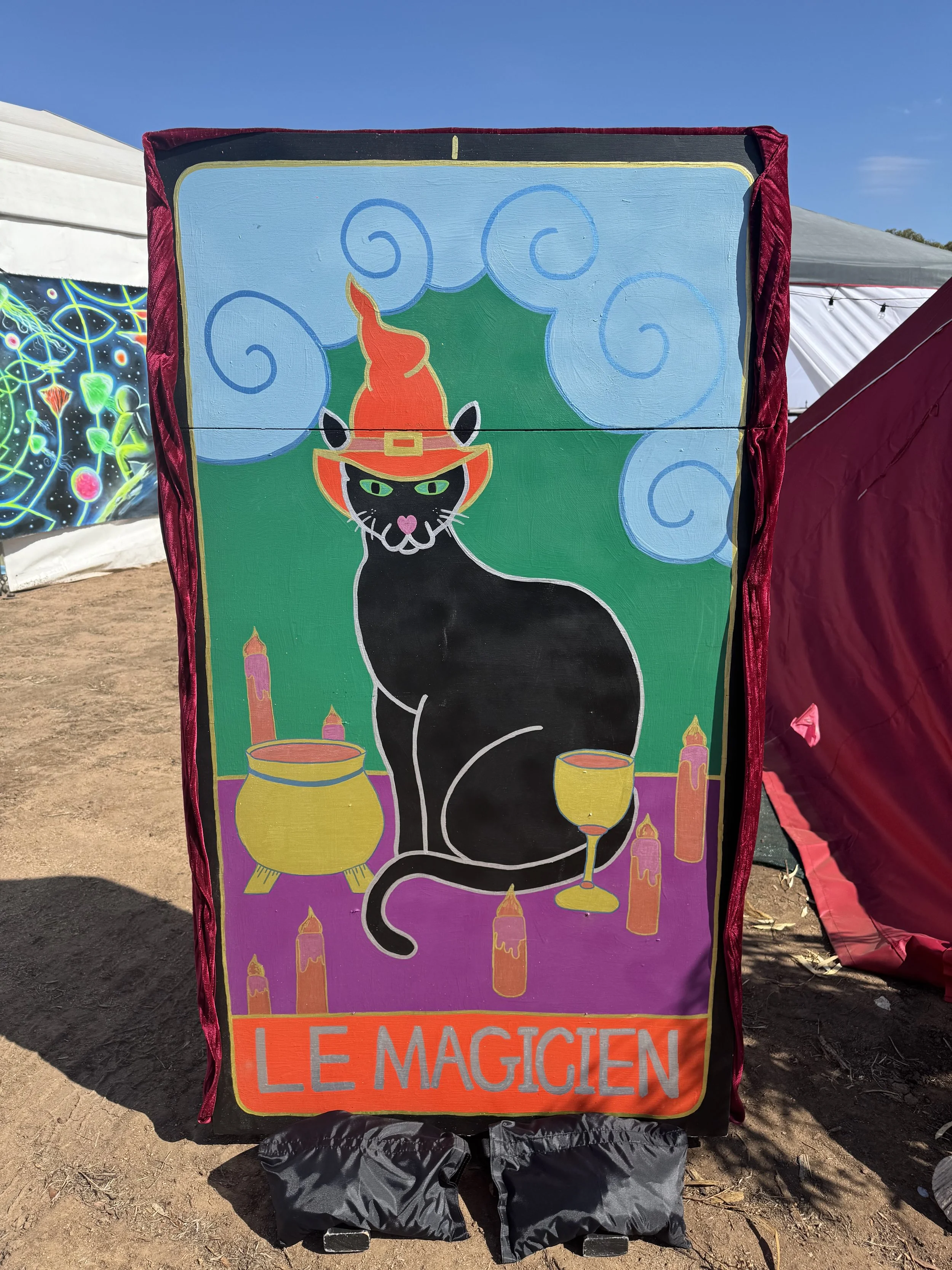 A colorful, illustrated sign featuring a black cat wearing a red wizard hat, with green eyes, surrounded by candles, a cauldron, and a wine glass, with the text "Le Magicien" at the bottom.