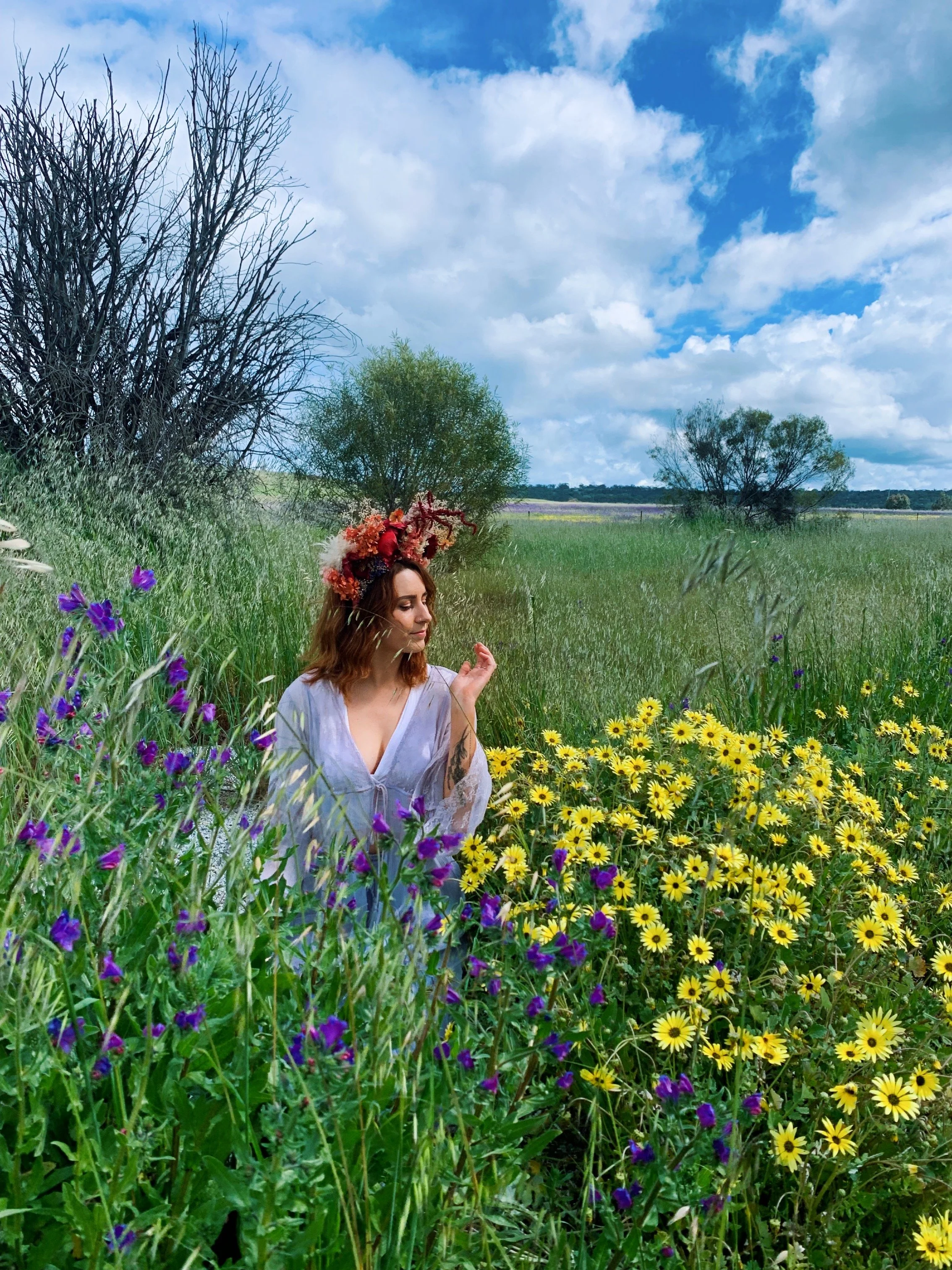 Woman in a white dress with a flower crown sitting in a field of yellow and purple wildflowers under a blue partly cloudy sky.