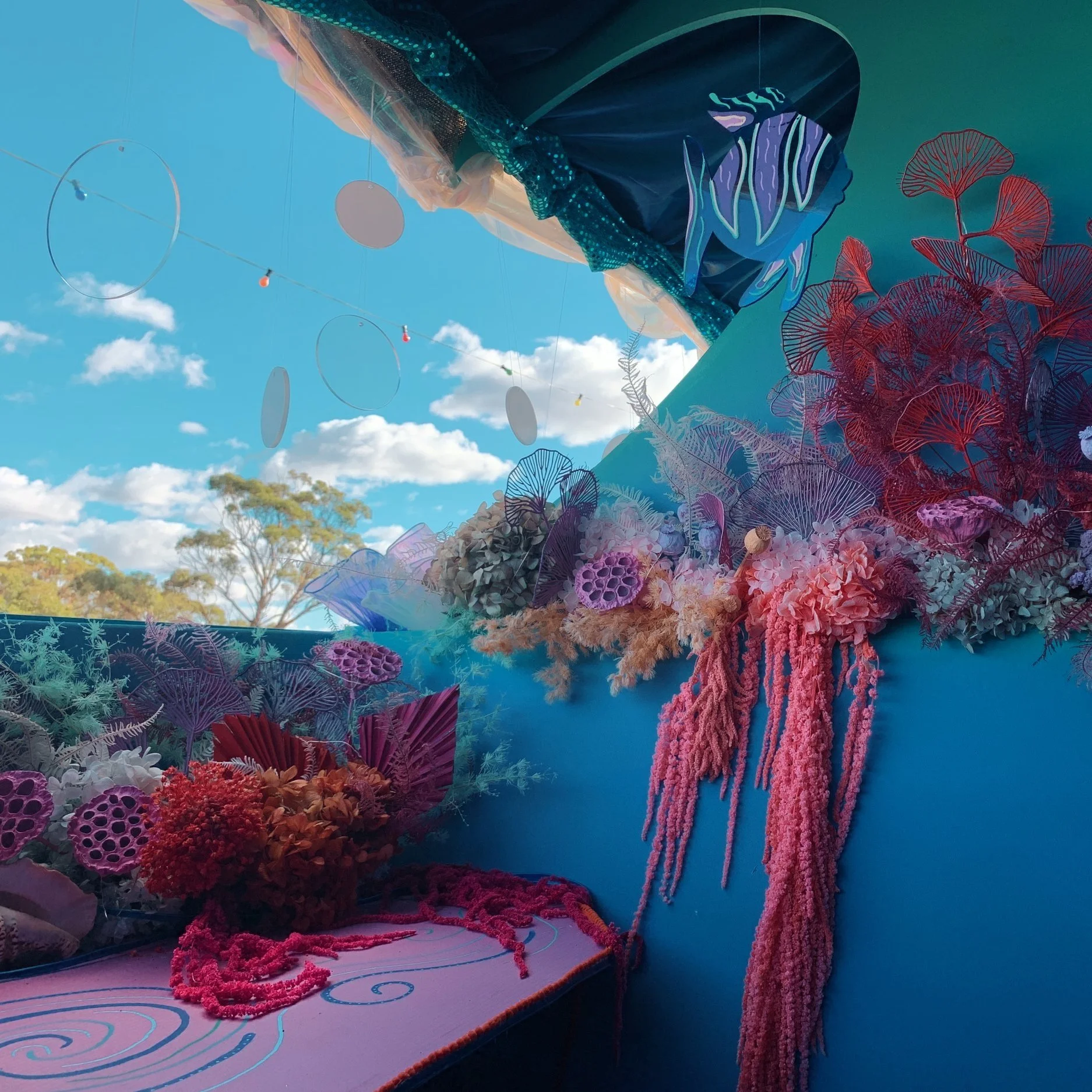 Colorful ocean-themed floral display with pink, purple, blue, and red coral and seaweed decorations against a bright blue sky with clouds.