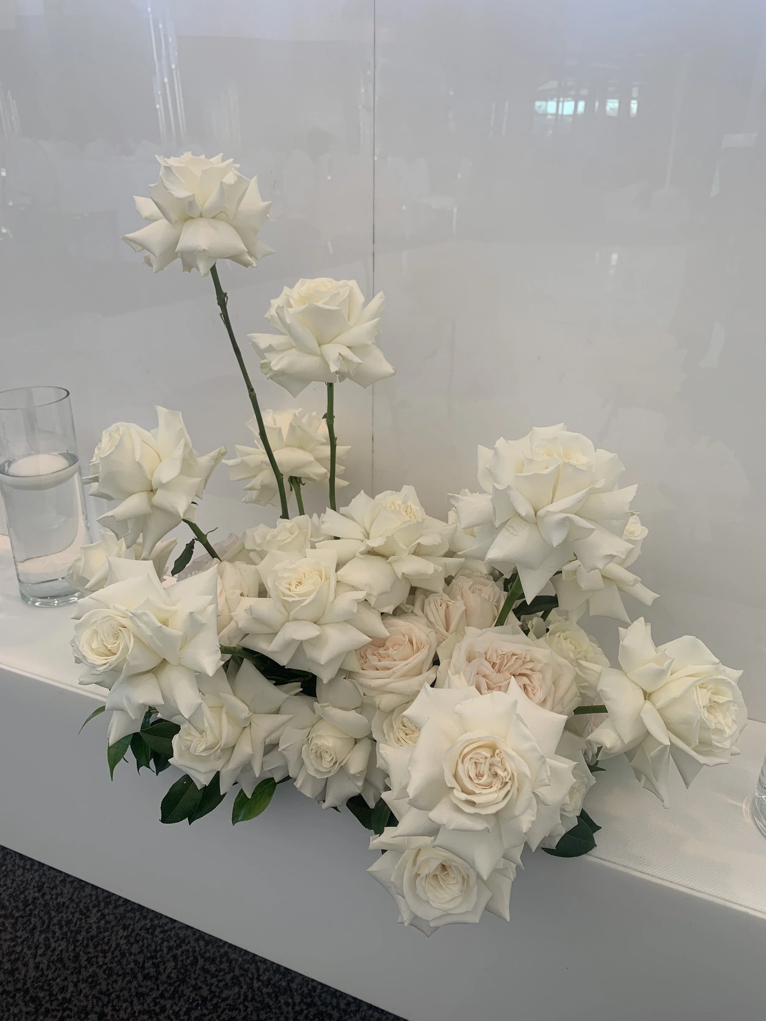 White flower arrangement with roses and lilies on a white surface, with two lit candles and a glass of water nearby.