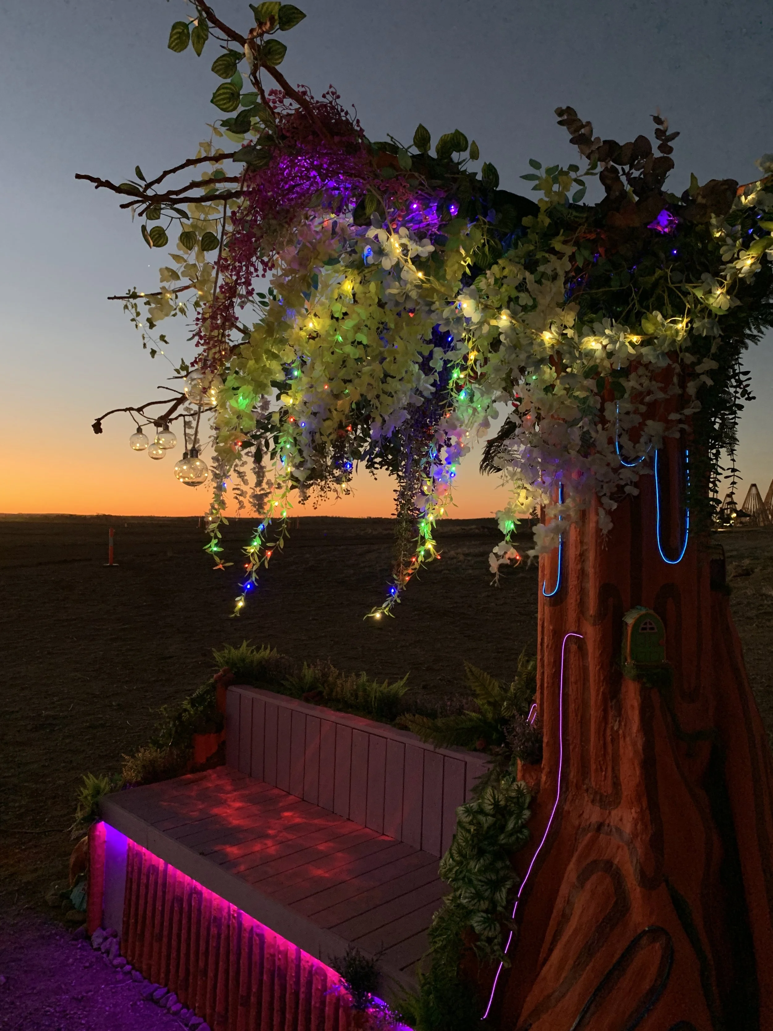 Decorated outdoor tree at sunset with colorful string lights, hanging ornaments, and a small wooden bench underneath.